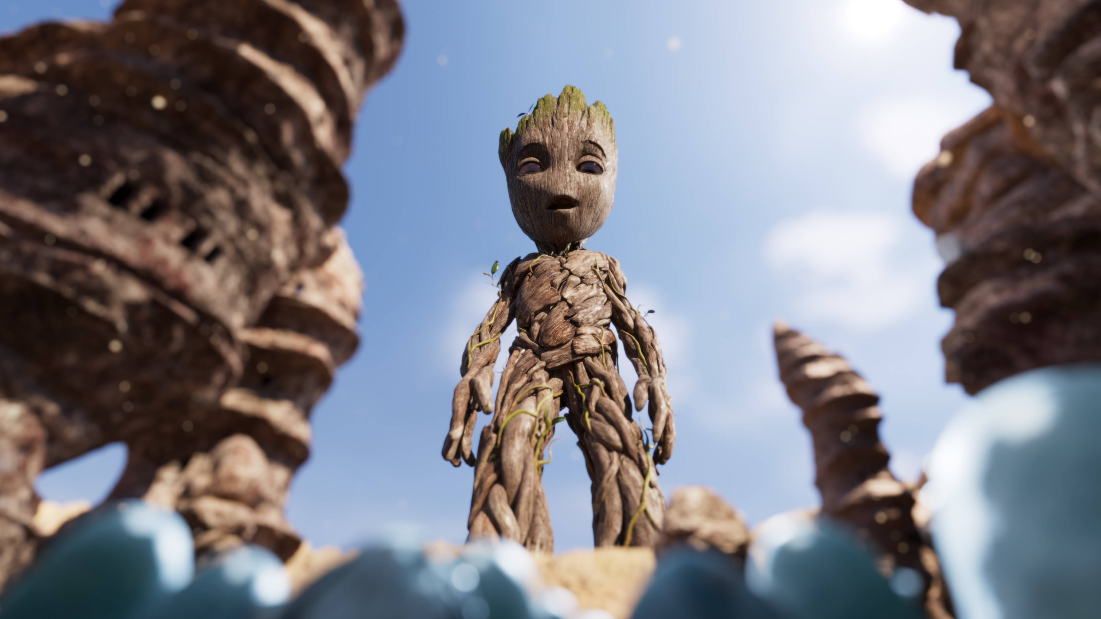 Baby Groot站立低角度 壁紙 — 一個細小嘅樹狀生物企喺巨大岩石形成物之間，喺有散雲嘅明亮藍天下。