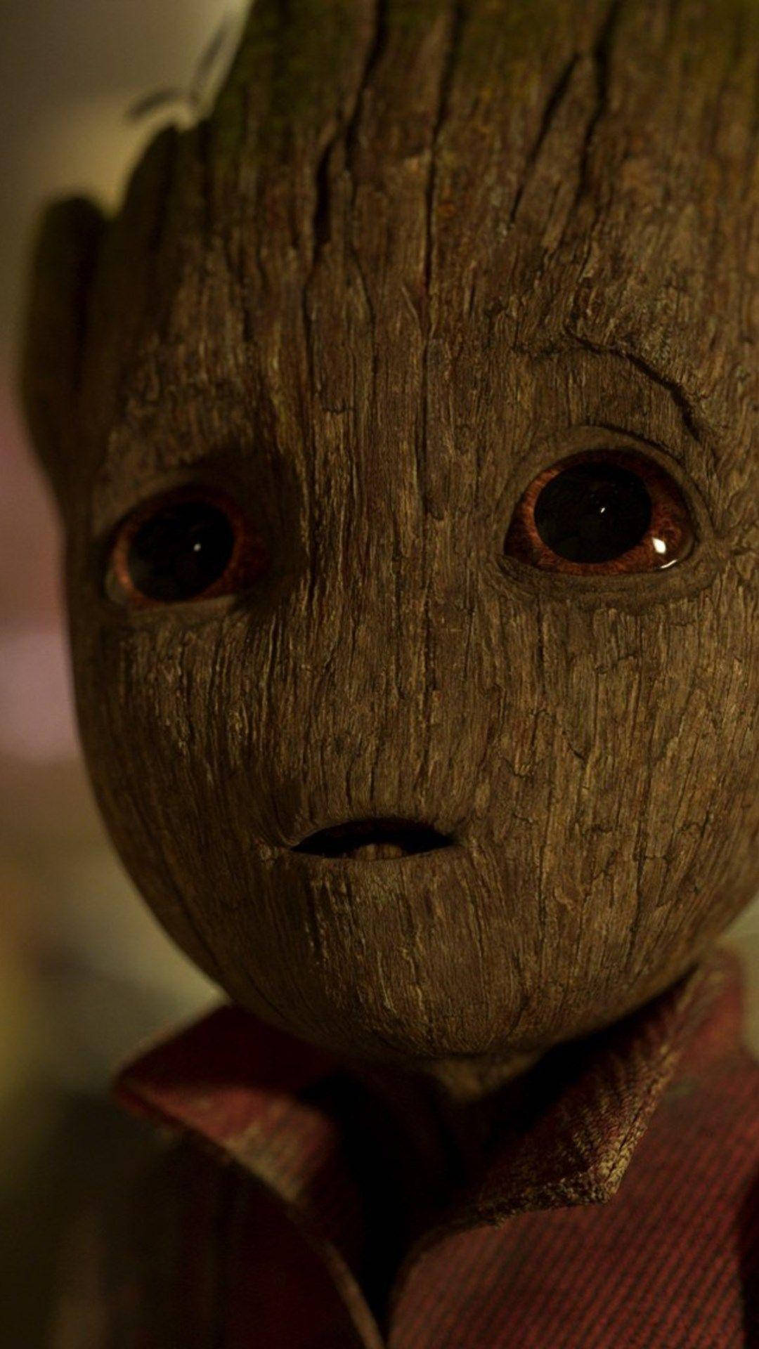 Baby Groot 臉部特寫 壁紙 — 近距離拍攝格魯特紋理木質臉部，有大黑眼睛和紅色有領襯衫，處於溫暖昏暗燈光下。