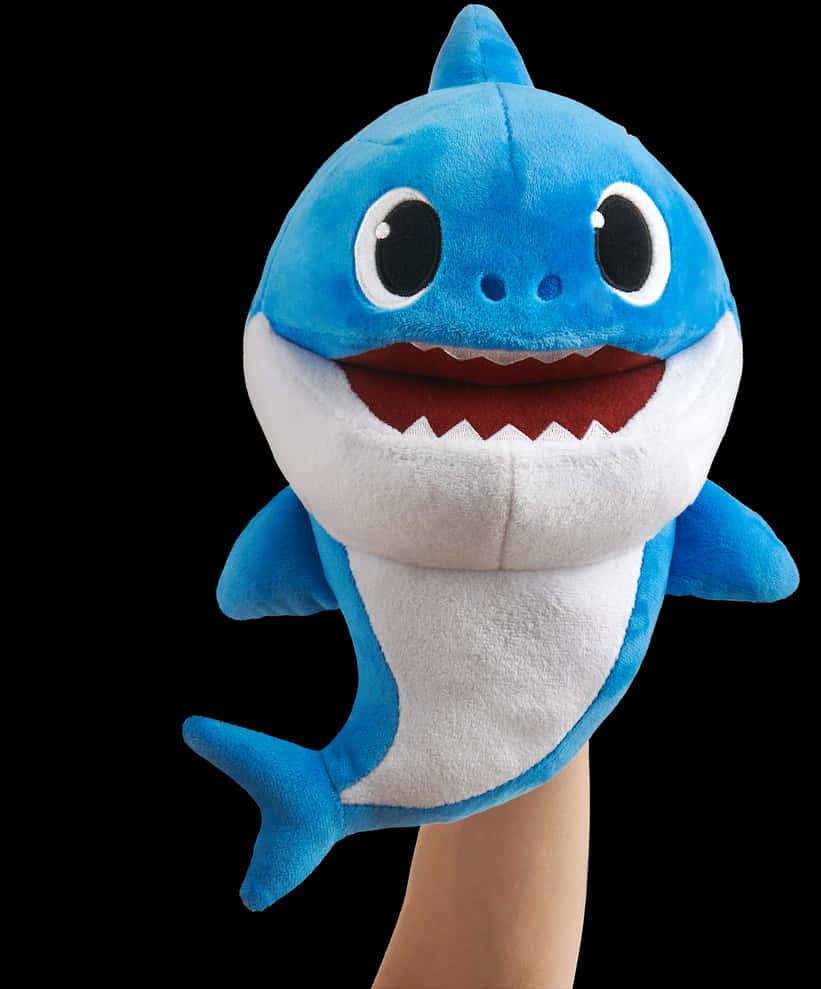 Baby Shark 毛絨玩具 壁紙 — 一隻藍白色毛絨嬰兒鯊魚手偶，有大眼睛和微笑的嘴巴，戴在手上，背景為黑色。