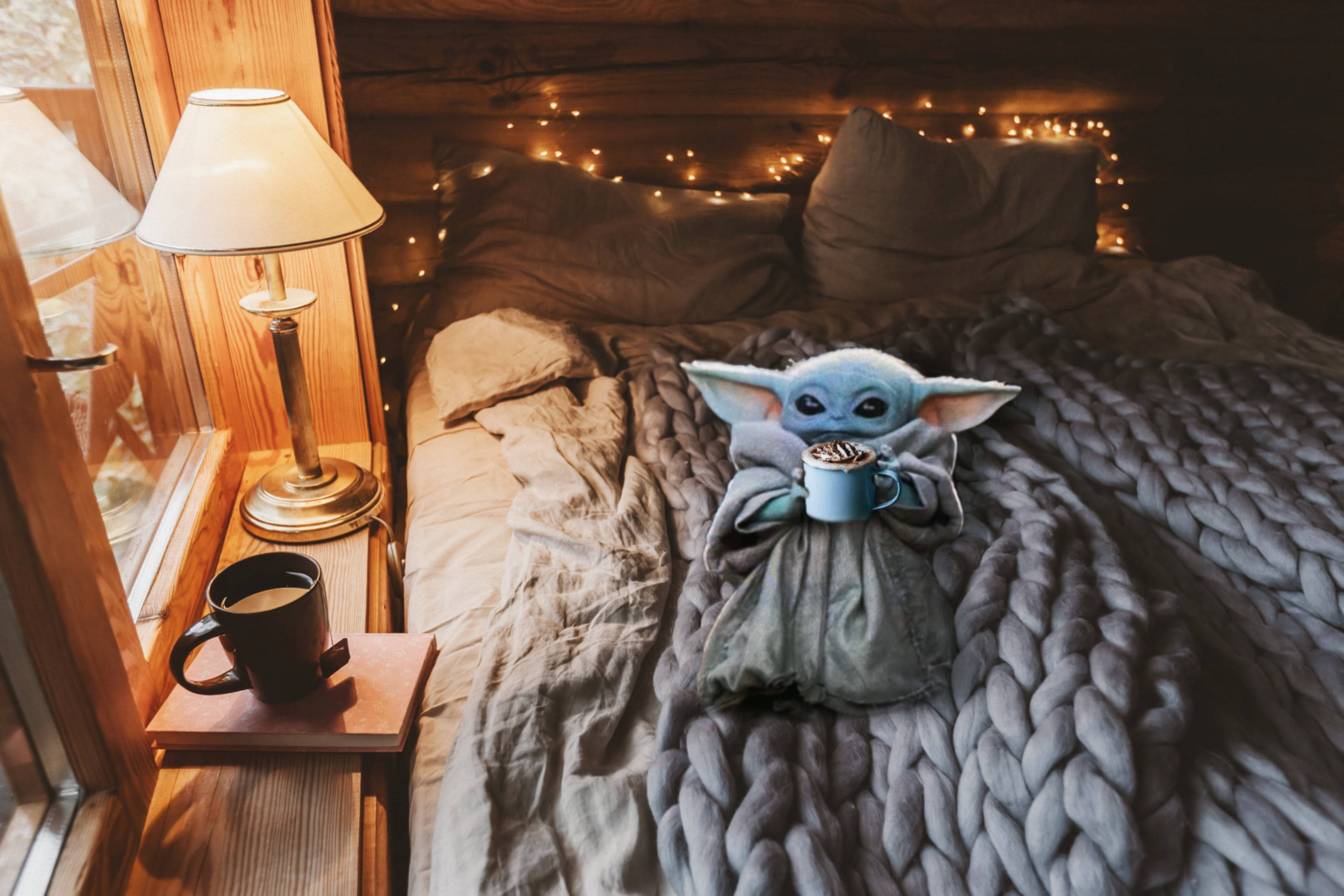 Baby Yoda 在溫馨冬日床鋪上 壁紙 — 一個藍色嘅Baby Yoda毛公仔舒適咁坐喺床上，喺溫暖嘅燈串同床頭燈下揸住一個杯。