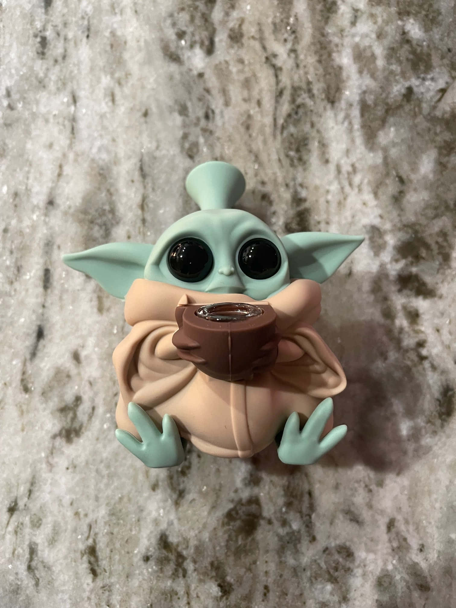 Baby Yoda展現新美學，可愛又時尚！ 壁紙 — 一個可愛的綠色尤達寶寶公仔，身穿米色長袍，在帶有斑點的灰色檯面上拿著一個棕色杯子。