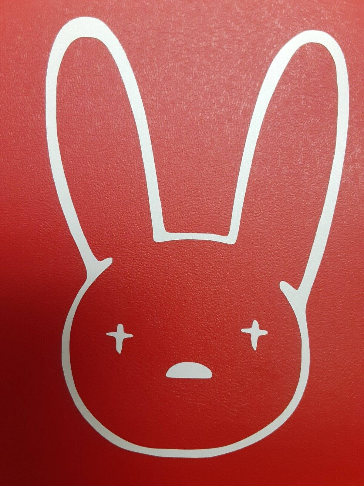 Bad Bunny 標誌 壁紙 — 在紋理紅色背景上，一個極簡主義的白色線條繪畫，描繪一個有交叉眼睛和小嘴巴的兔子臉。