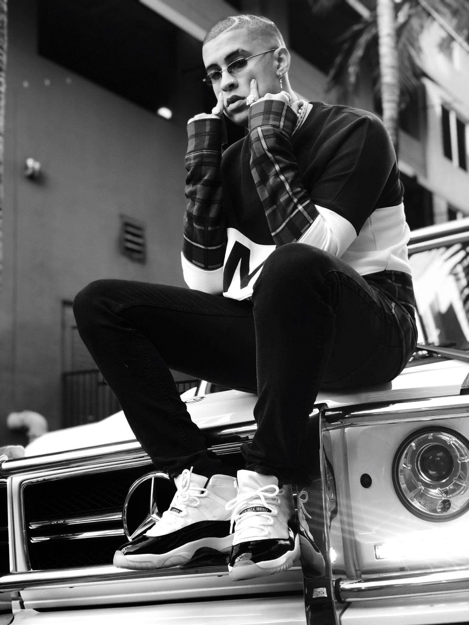 Bad Bunny 坐於車頂 壁紙 — 一位時尚男士戴著太陽眼鏡和格紋袖子，自信地坐在一輛經典汽車上，背景是單色城市場景。