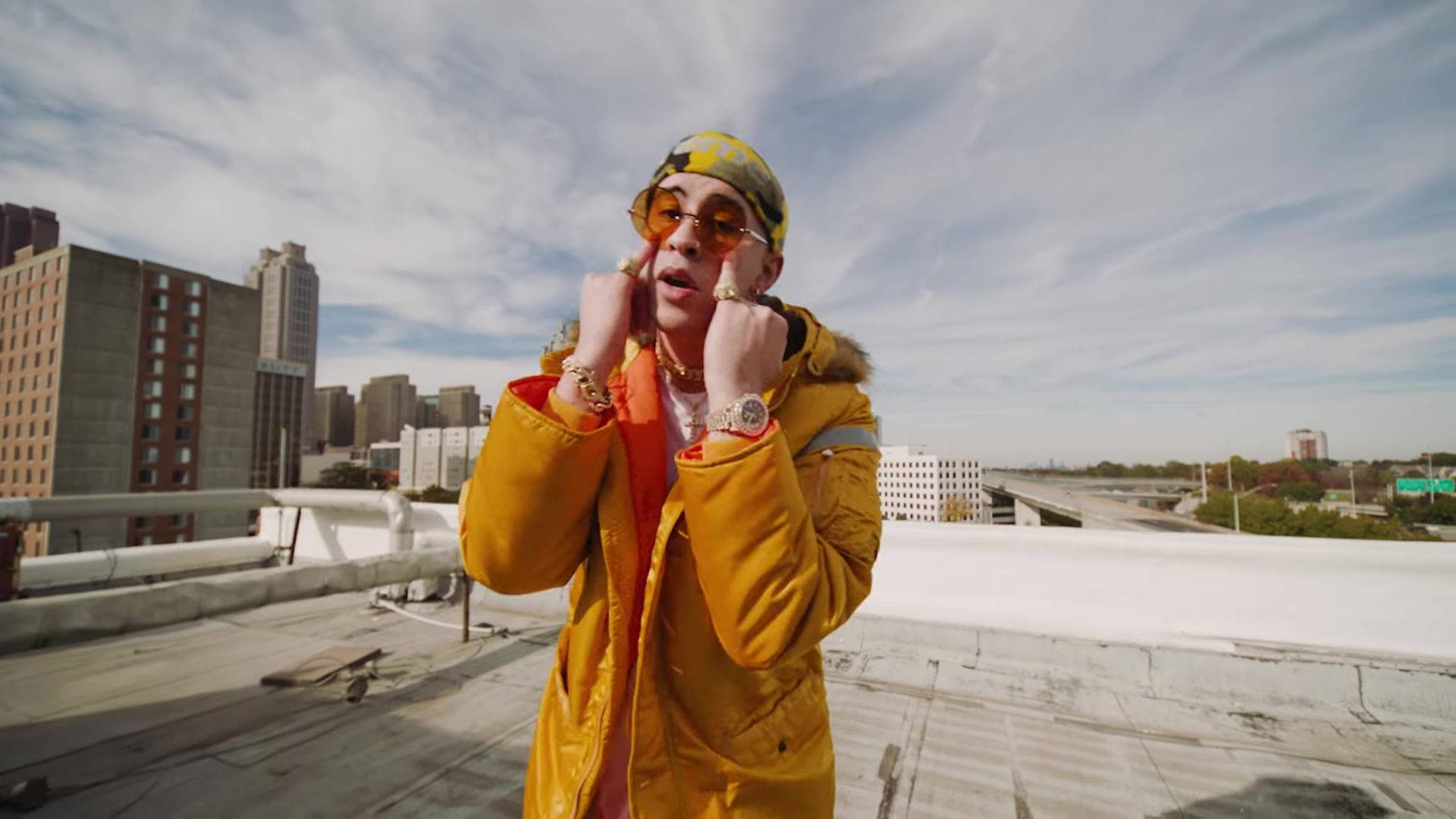 Bad Bunny 橙色服裝搭配 壁紙 — 一位時尚人士身穿亮黃色外套和圖案帽子，在部分多雲的天空下自信地在城市屋頂上擺姿勢。
