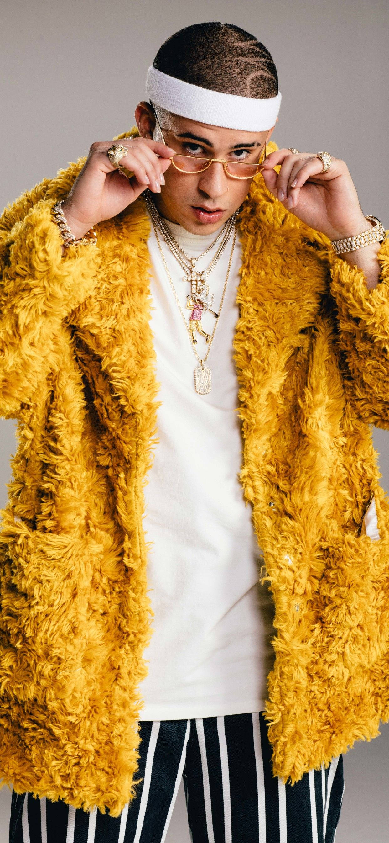 Bad Bunny 毛茸茸黃色外套 壁紙 — 一位時尚男士穿著蓬鬆的黃色外套，在灰色中性背景前調整眼鏡。