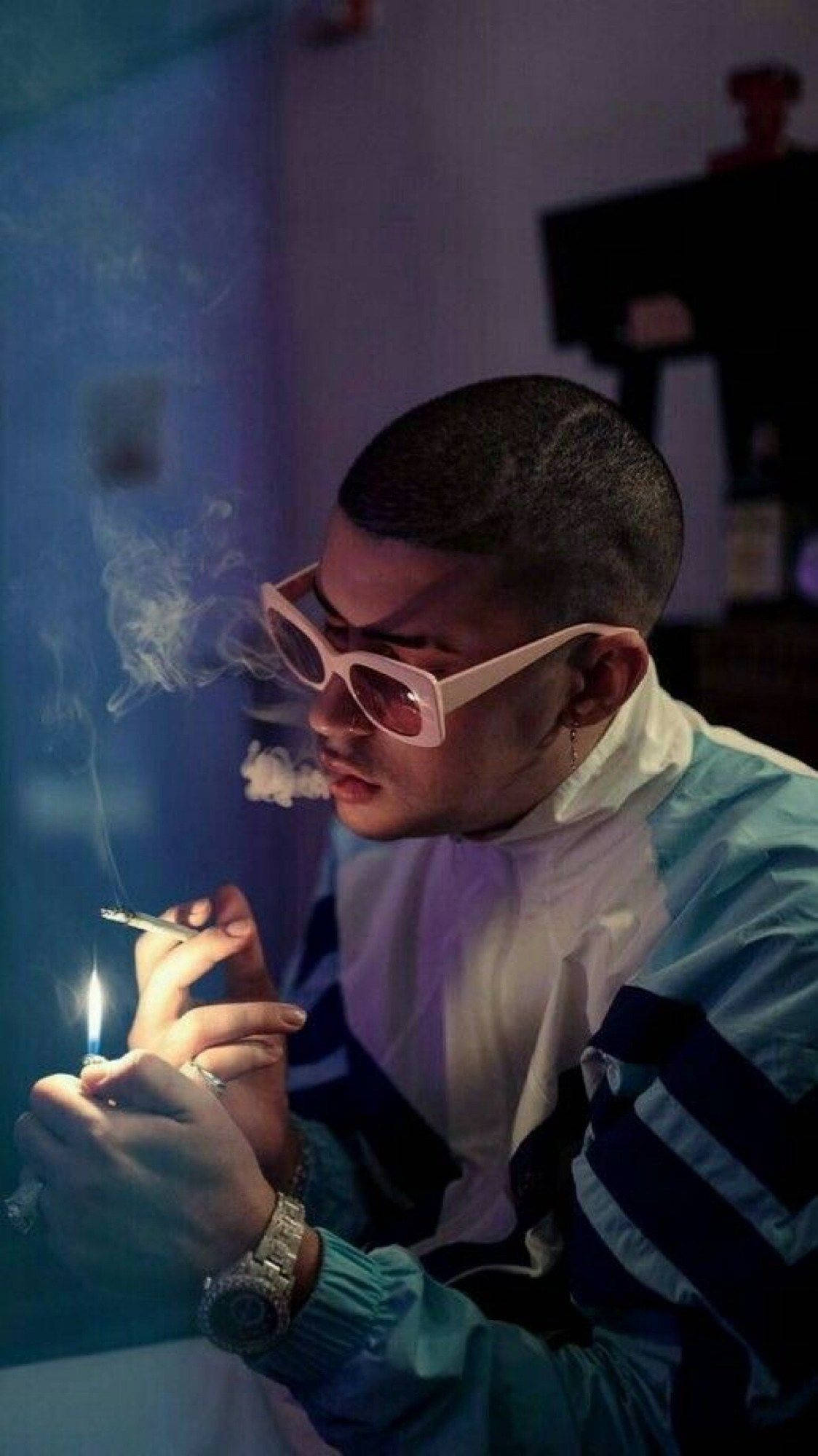 Bad Bunny 煙草壁紙 壁紙 — 一名戴著粉紅色太陽眼鏡的男子在昏暗的藍色燈光房間裡點燃香煙，煙霧在他臉龐周圍繚繞。