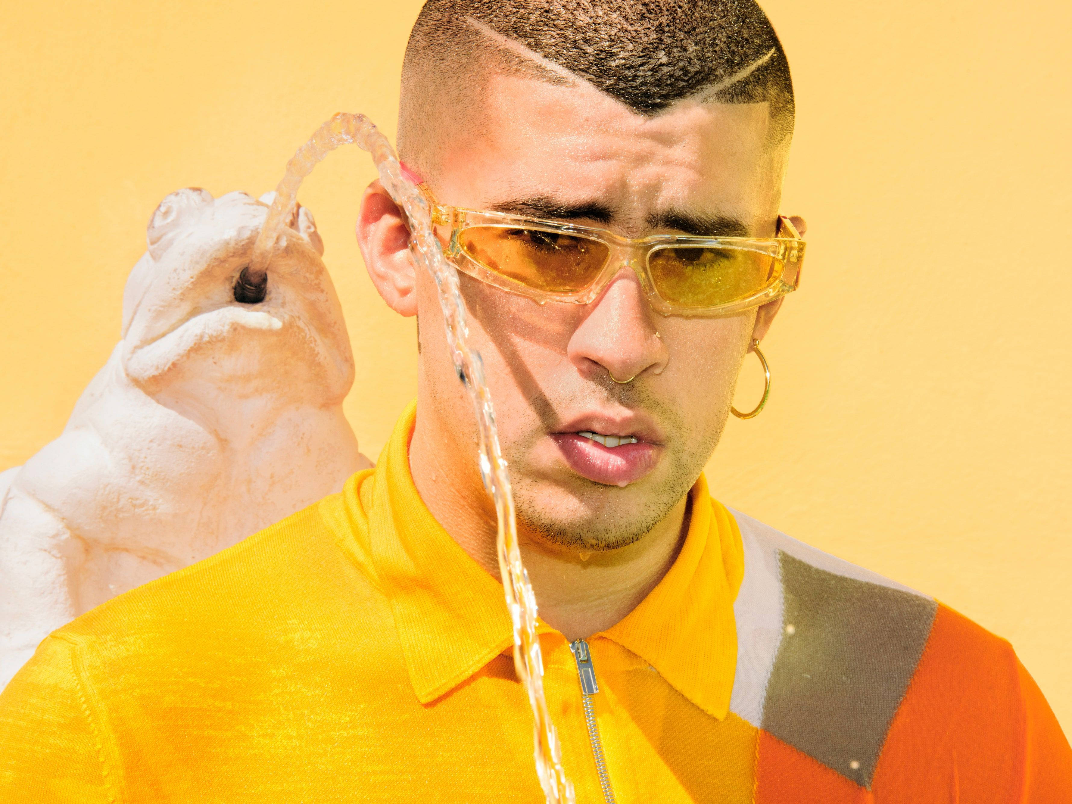 Bad Bunny 白色青蛙噴泉 壁紙 — 一個戴著黃色太陽眼鏡和穿著亮黃色襯衫的男人站著，身後一個白色青蛙雕像噴出水柱，背景是純黃色。