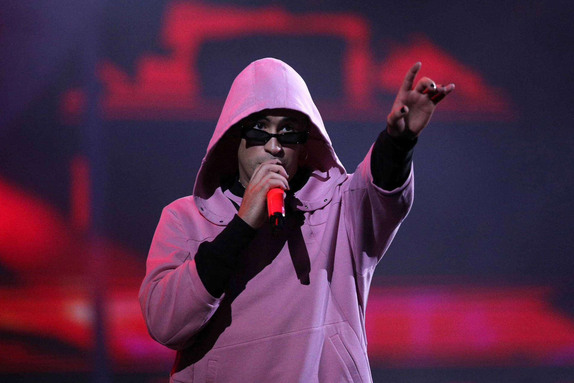 Bad Bunny 穿紫色連帽衫 壁紙 — 一位表演者穿著粉紅色連帽衫和黑色太陽眼鏡，在鮮紅色的舞台燈光下對著紅色麥克風唱歌。