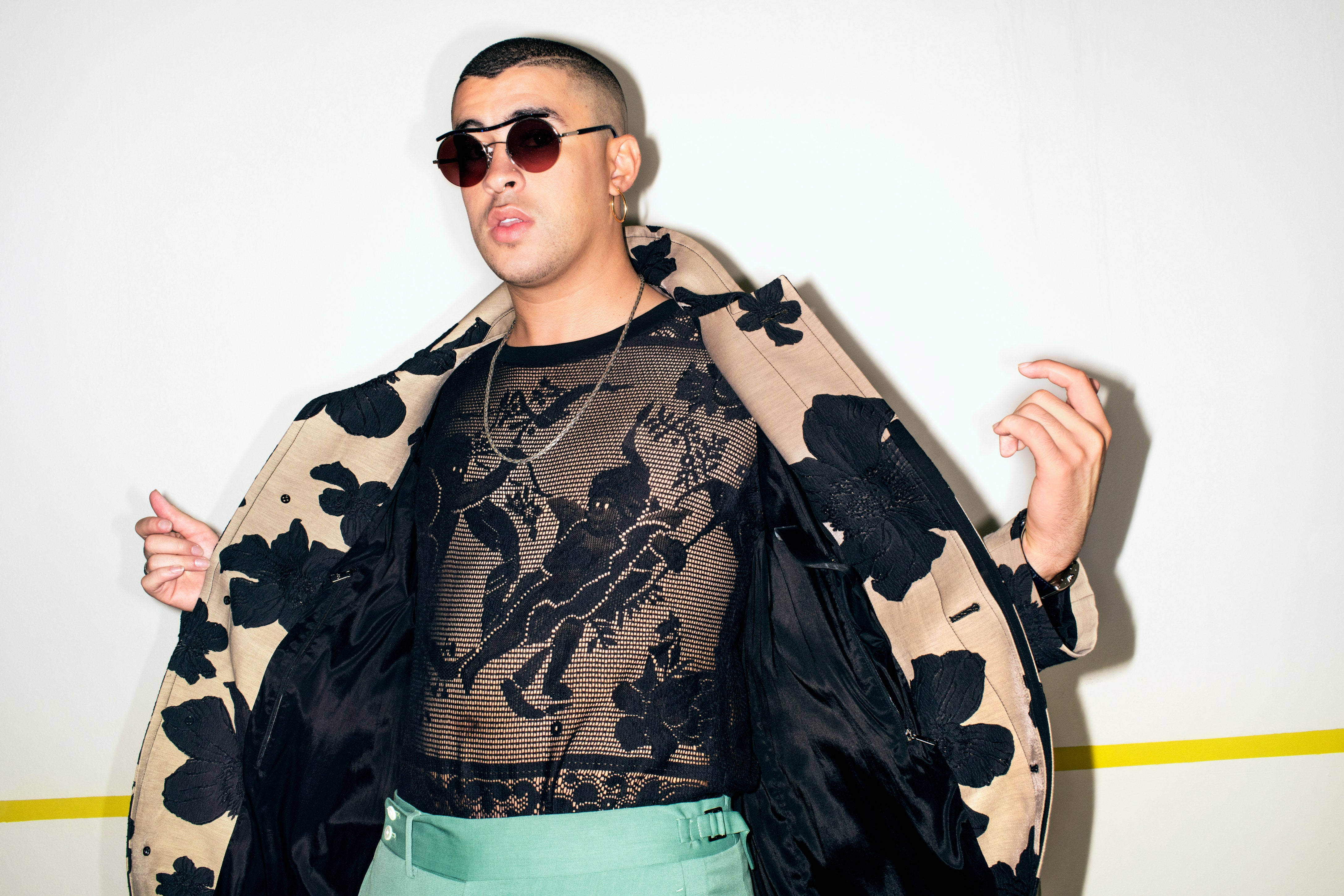 Bad Bunny 穿透視上衣 壁紙 — 一位時尚男士戴著太陽眼鏡和花卉圖案外套，自信地站在帶有黃色條紋的純白牆前擺姿勢。