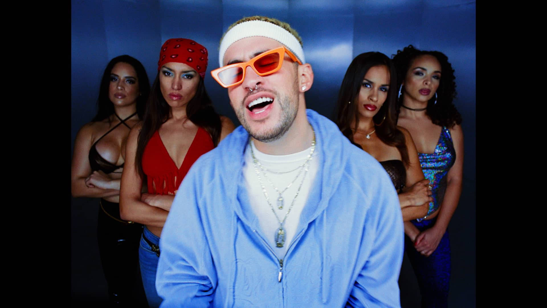 Bad Bunny 與四位女生同框 壁紙 — 一名戴橙色太陽眼鏡和藍色連帽衫的男子大笑，身後有四名穿著暴露的女子站在藍色牆前。