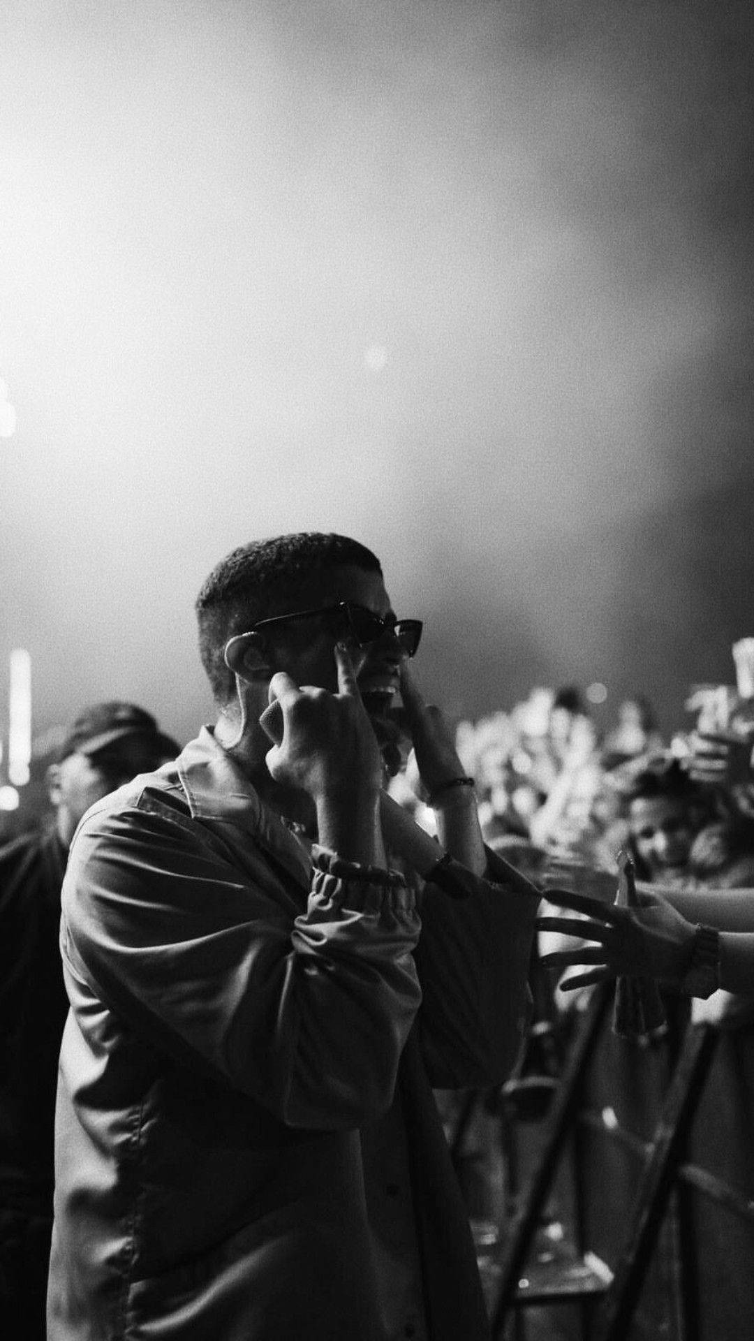 Bad Bunny 與粉絲互動 壁紙 — 一名戴太陽眼鏡的男子在明亮的舞台燈光下向音樂會人群做手勢。