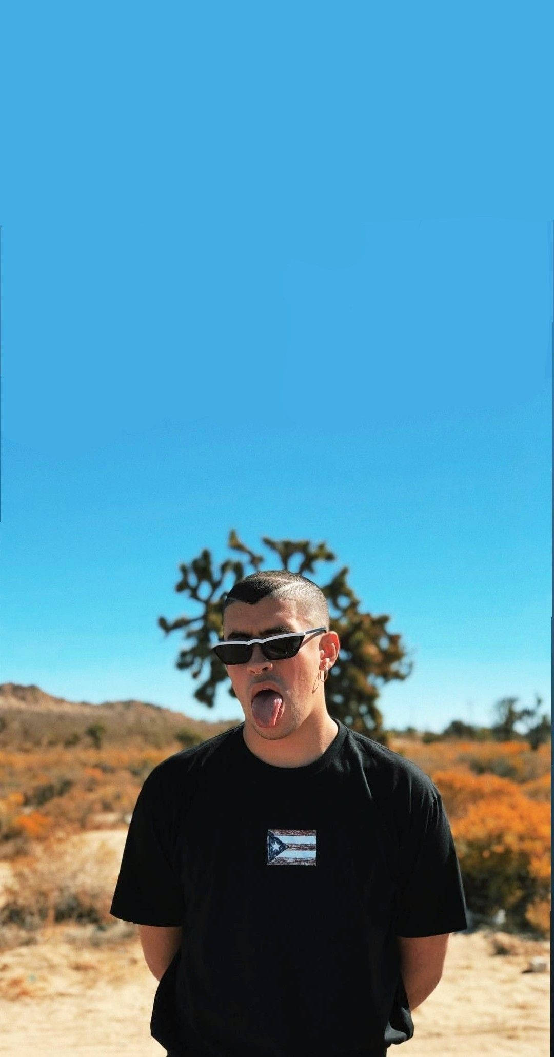 Bad Bunny 舔舌表情 壁紙 — 一個戴著太陽眼鏡的年輕男子在廣闊的藍天下伸出舌頭，下方有約書亞樹和沙漠灌木。