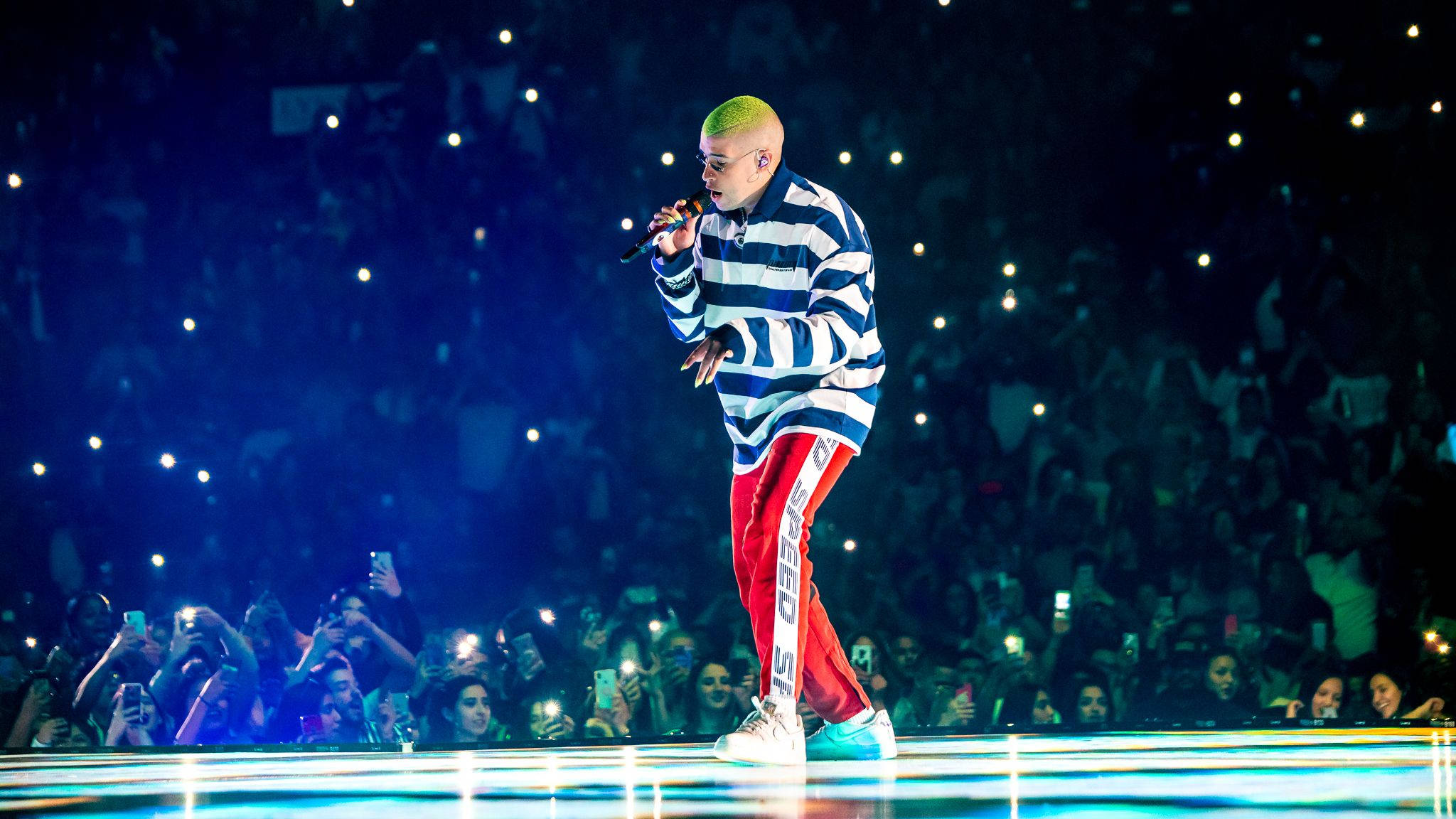 Bad Bunny 舞台前與粉絲同框 壁紙 — 一位表演者擁有螢光綠色頭髮，在發光的舞台上對著咪高峰唱歌，粉絲們在藍色燈光下舉起手機。