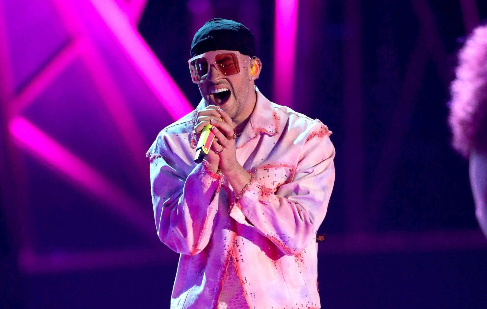 Bad Bunny 舞台演出壁紙 壁紙 — 一位男性表演者戴著粉紅色太陽眼鏡和一件破舊的粉紅色外套，在充滿活力的紫色舞台燈光下熱情地對著麥克風唱歌。