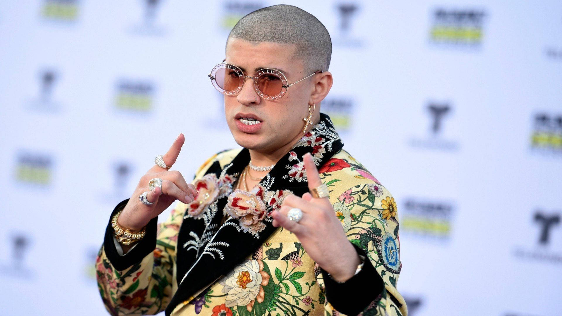 Bad Bunny 花紋外套 壁紙 — 一名光頭男子戴著圓形太陽眼鏡，穿著鮮豔的花卉外套，在記者會上自信地做手勢。