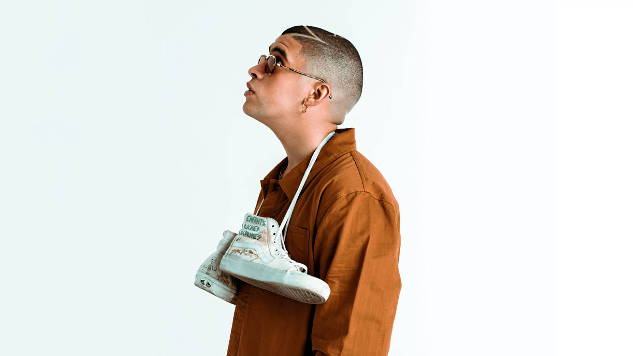 Bad Bunny 頸掛白色 Vans 鞋 壁紙 — 一個穿著橙色襯衫的時尚人士，手持白色運動鞋，仰望純色淺色背景。