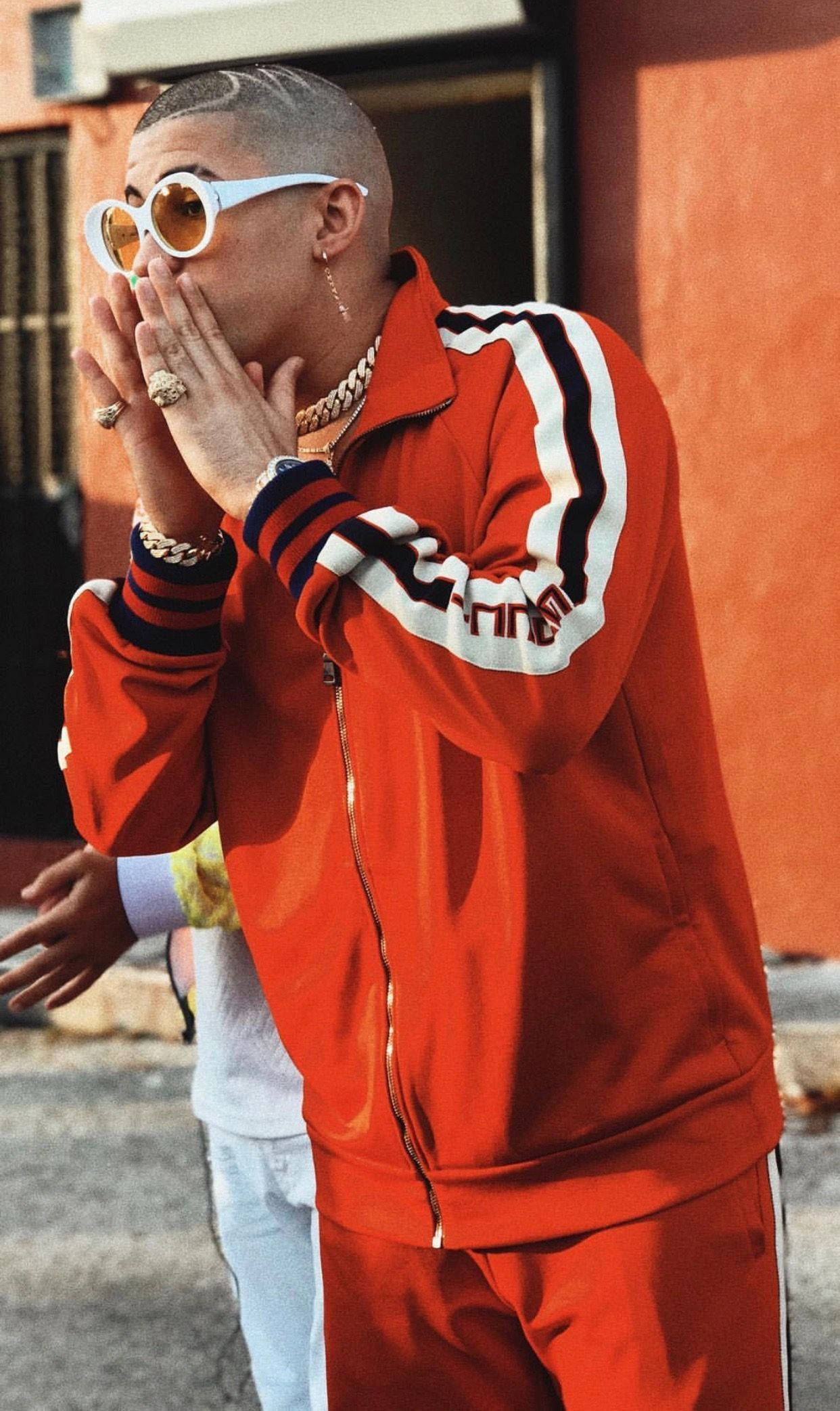 Bad Bunny 驚訝表情 壁紙 — 一位時尚男士身穿大膽的紅色運動服和白色太陽眼鏡，雙手捂住嘴巴，站在暖色調的城市牆壁前。