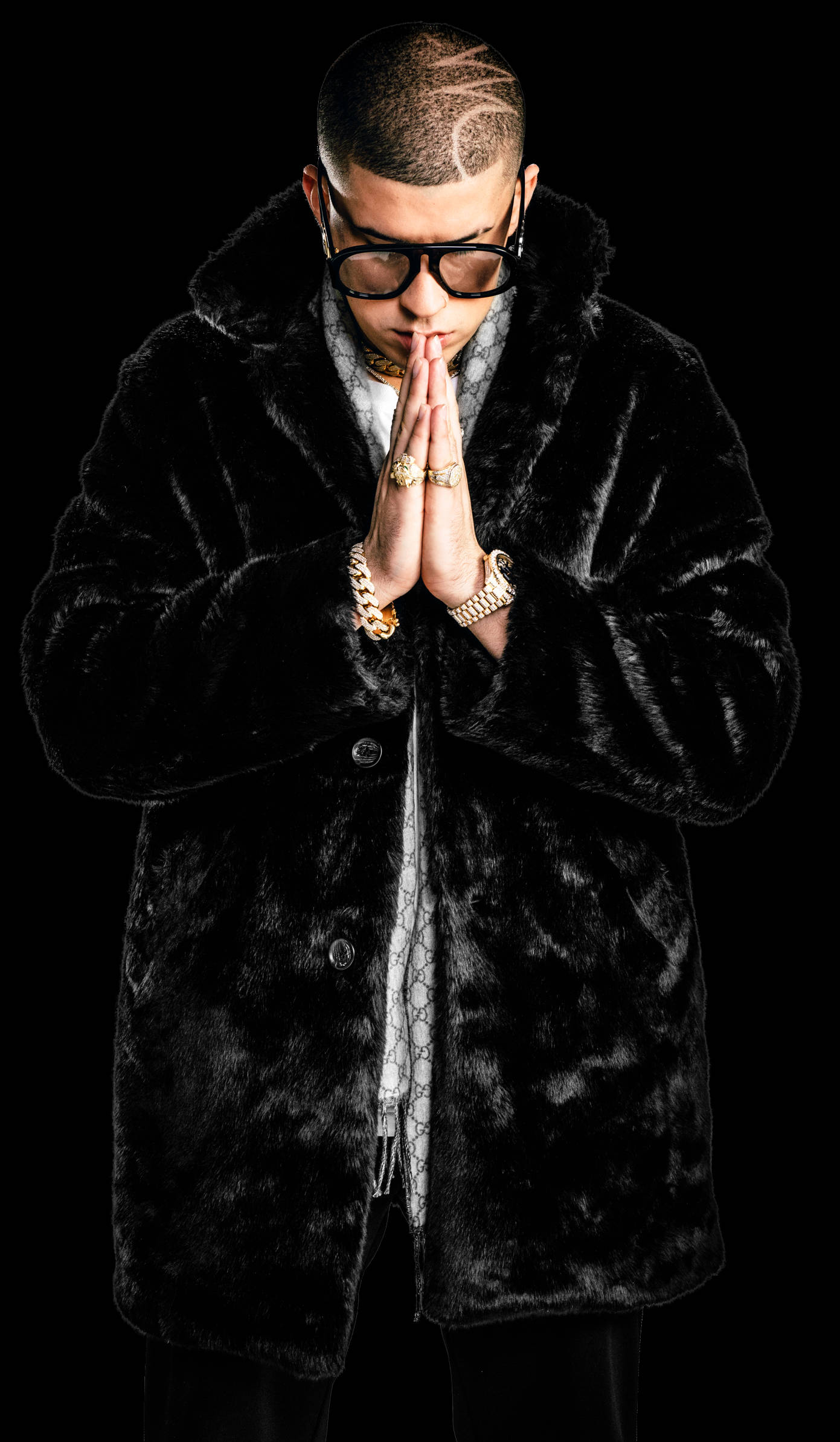 Bad Bunny 高光黑色毛皮 壁紙 — 一個穿著黑色毛皮大衣和眼鏡的男人在黑色背景前合十祈禱。