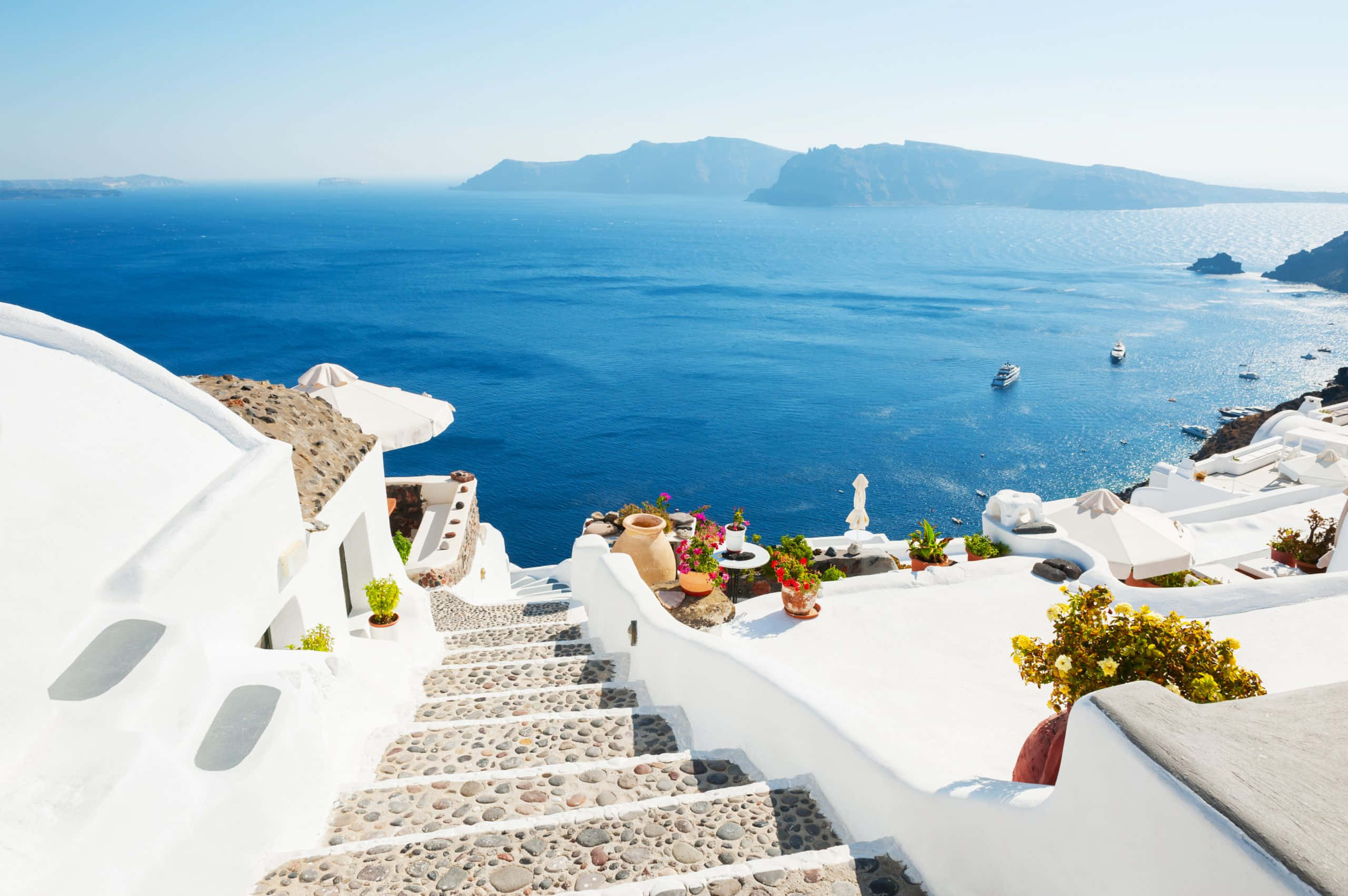 Greek Beach 桌布 壁紙 — 白色粉刷的樓梯通向充滿活力的藍色愛琴海，遠處有火山懸崖，天空晴朗。