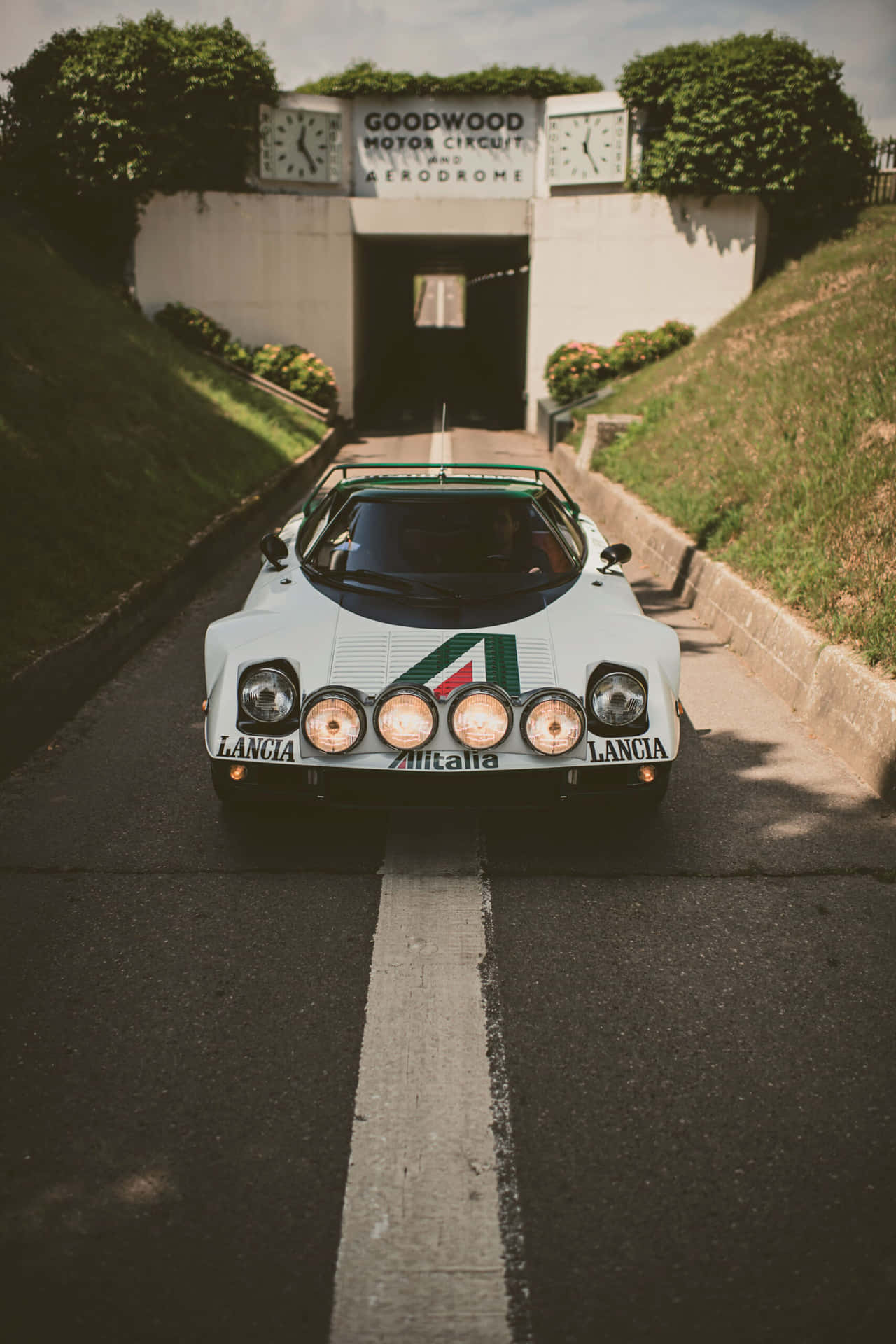  壁紙 — 一輛白色Lancia 037拉力賽車，帶有綠色點綴和四個圓形頭燈，在通往古德伍德賽道隧道的瀝青路上向鏡頭駛來。