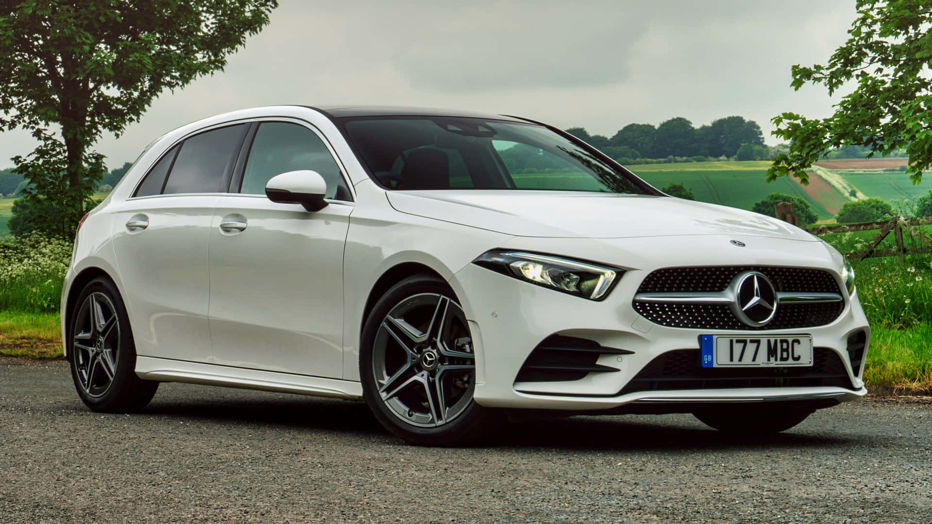 Mercedes Benz A-class 桌布 壁紙 — 一架時尚的白色 Mercedes-Benz A-Class 掀背車停泊在碎石路上，背景是綠色的田野和樹木，天空多雲。