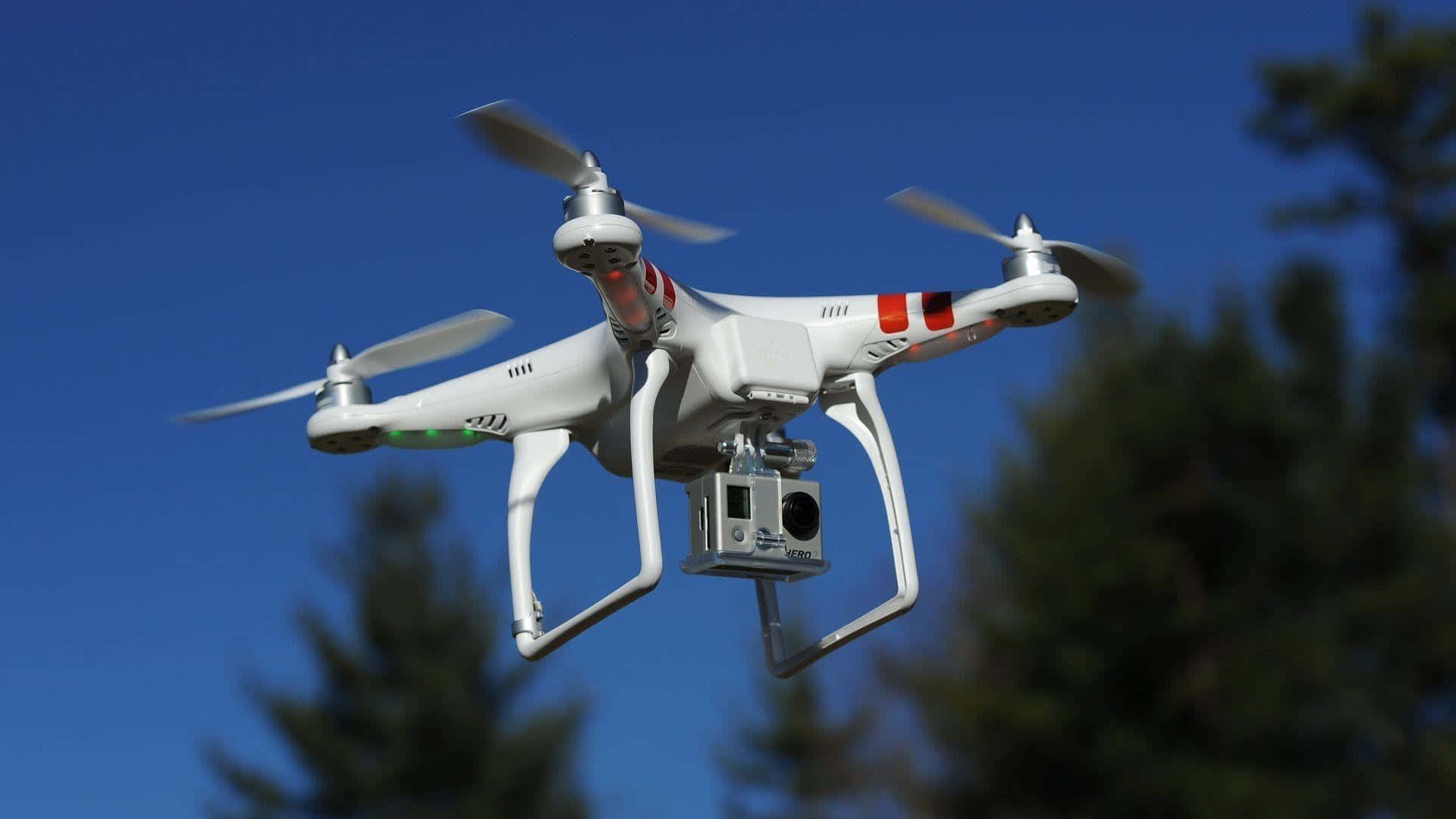 Drone Camera 桌布 壁紙 — 一架白色四軸無人機帶旋轉槳葉，在清晰藍天中盤旋，下方有模糊的綠色樹木。