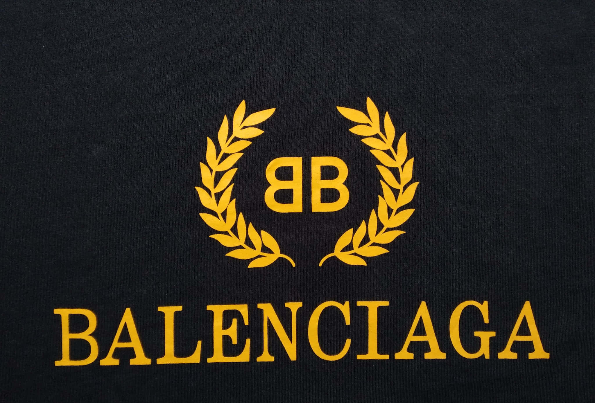 踏入Balenciaga的時尚世界 壁紙 — 大膽金色 Balenciaga 標誌配月桂花環，特寫於紋理黑色布料上。
