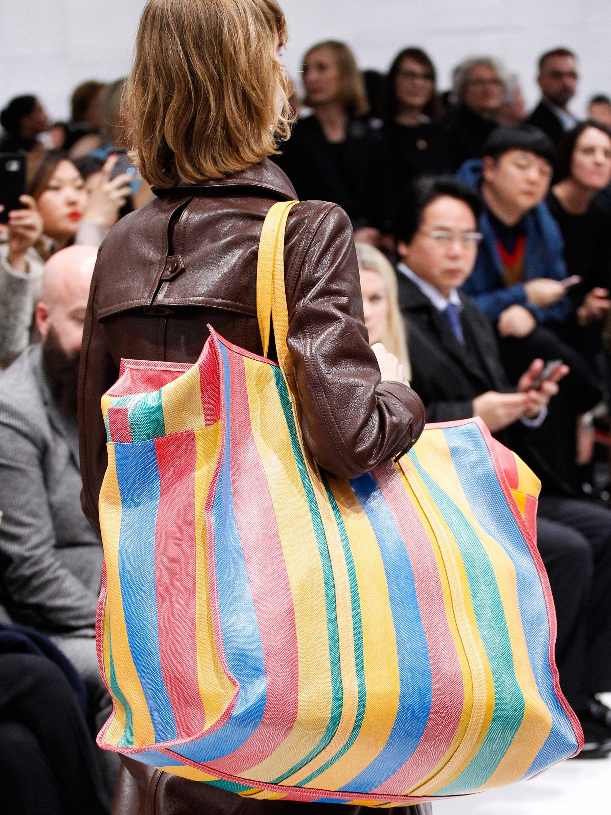 Balenciaga 泰式手袋 壁紙 — 一位身穿棕色皮褸的女性，在觀眾注視下，拿著一個色彩繽紛的條紋大手提袋走過天橋。