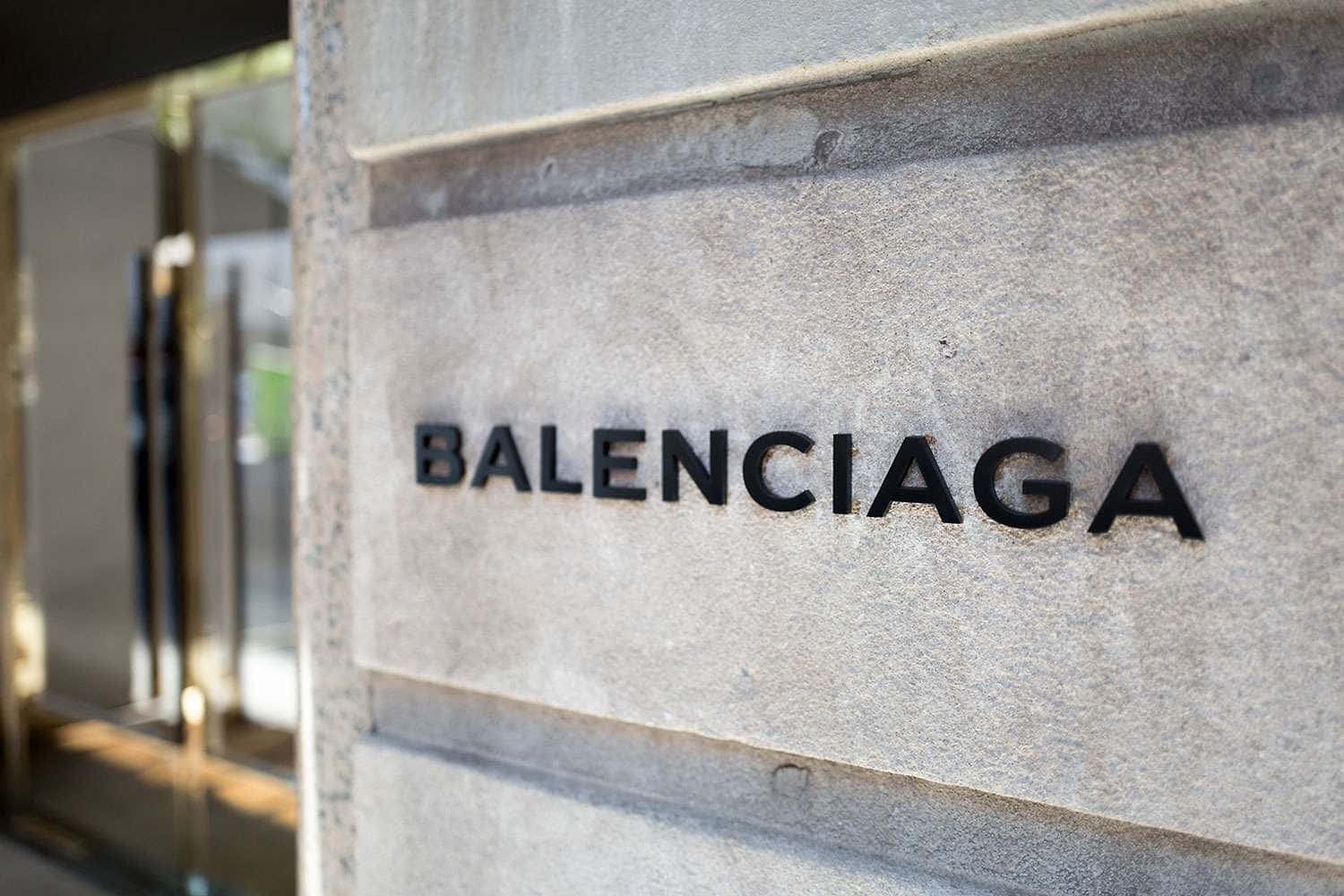 以 Balenciaga 保持時尚領先 壁紙 — 特寫：光滑黑色Balenciaga標誌嵌入紋理灰色石牆，背景有玻璃門。