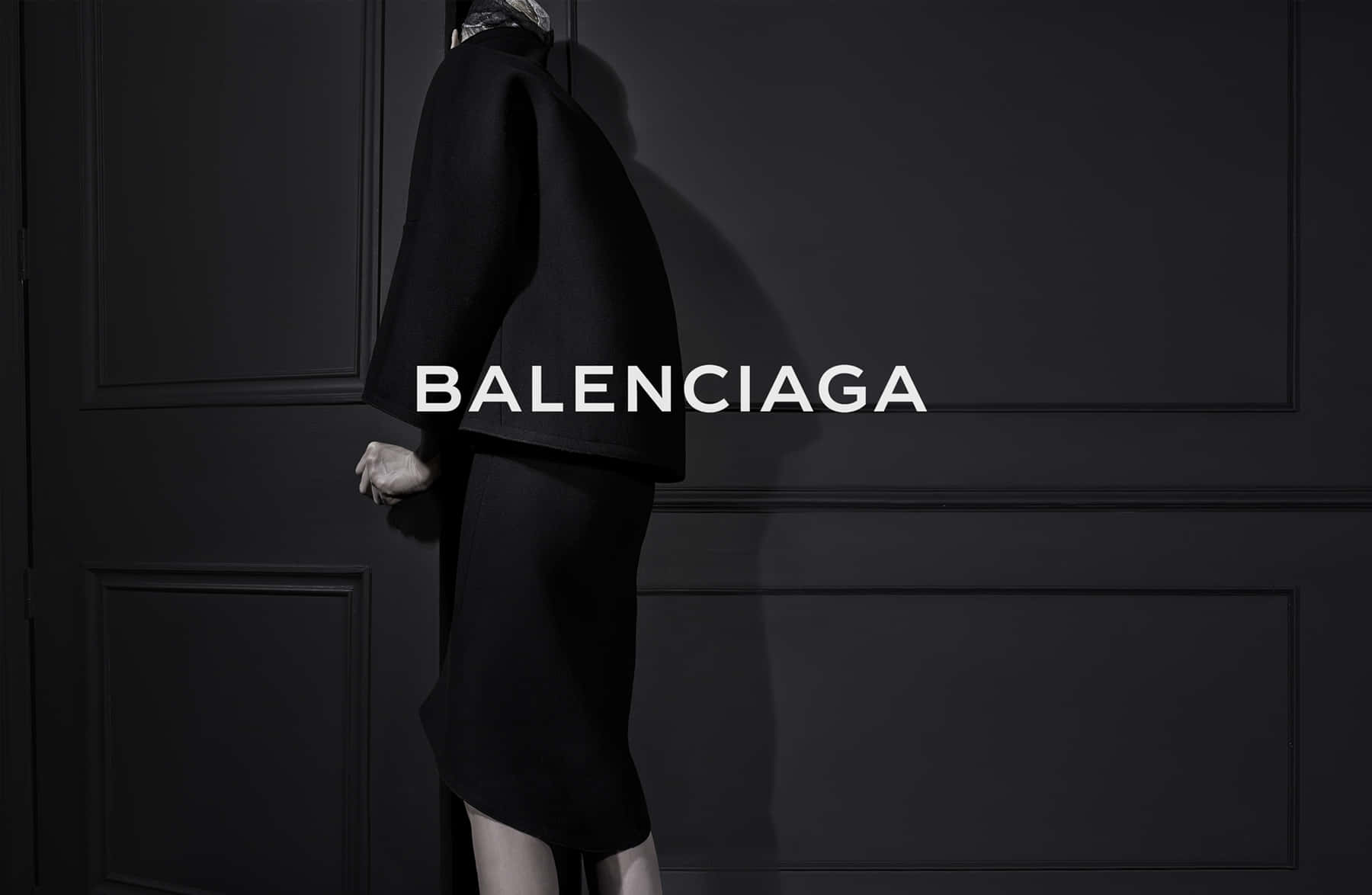 打破規則，展現Balenciaga的時尚宣言 壁紙 — 一個穿著黑色Balenciaga服裝的人側面站立在深色鑲板牆前，品牌名稱顯眼地展示。