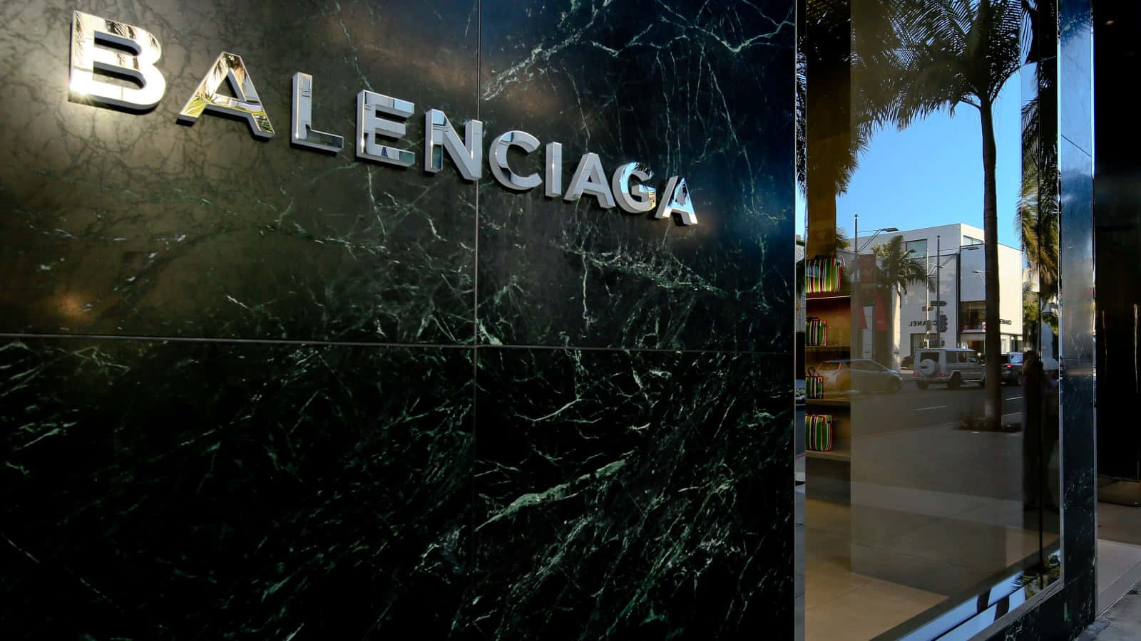 以Balenciaga展現個人風格 壁紙 — 閃亮的鉻色 Balenciaga 標誌在深綠色大理石外牆上閃耀，旁邊是玻璃店面，映照著棕櫚樹和城市街道。
