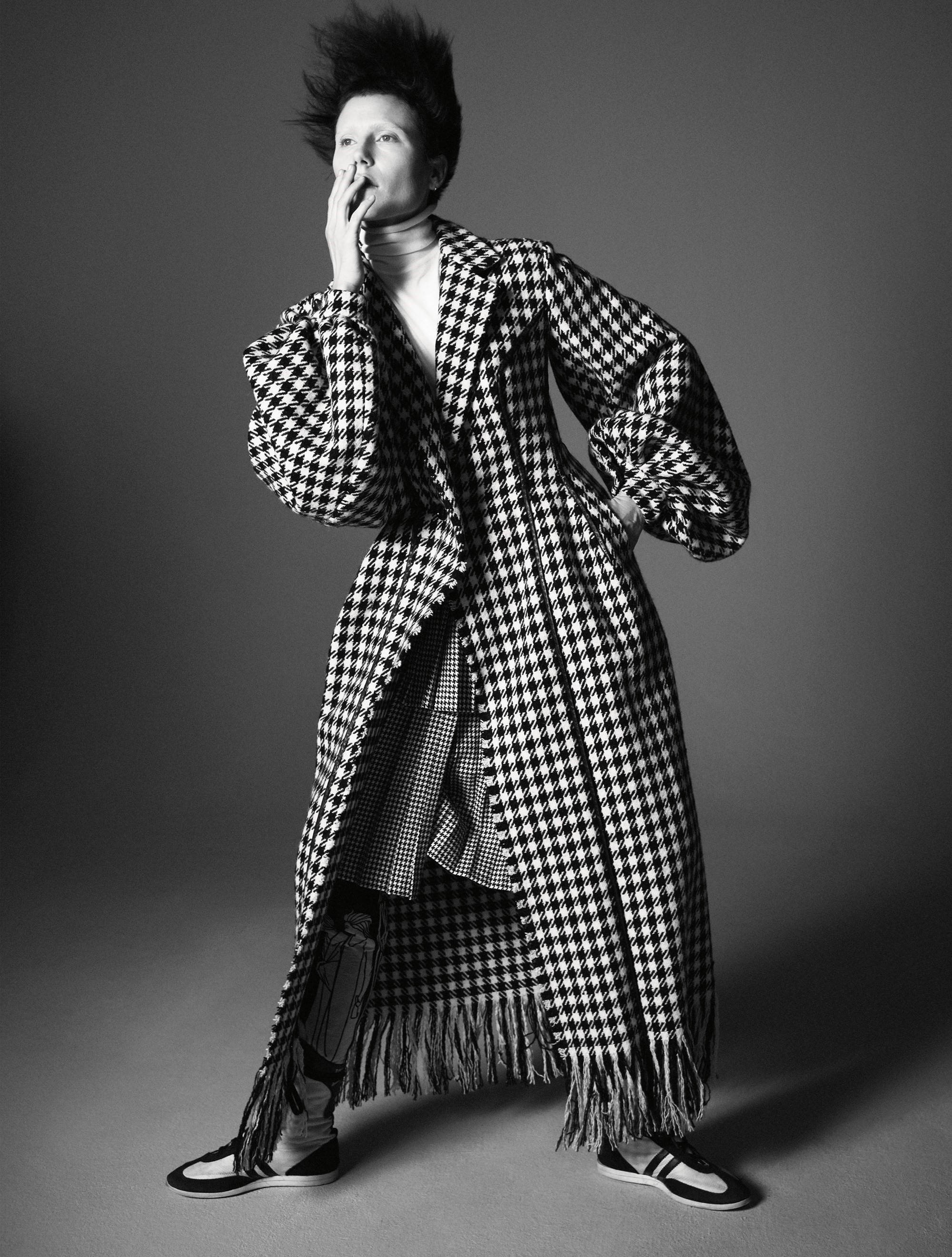 Balenciaga 格紋外套 壁紙 — 一個人穿著戲劇性的千鳥格大衣，帶有流蘇下擺和超大袖子，在純灰色工作室背景前擺姿勢。