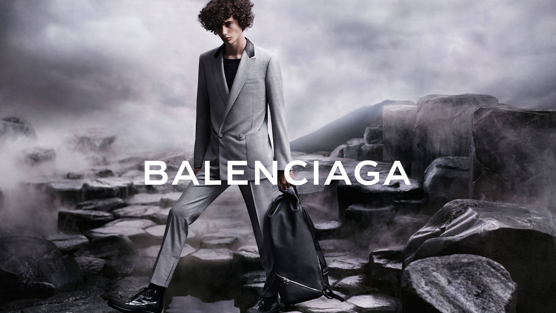 Balenciaga男模西裝 壁紙 — 一位身穿灰色西裝的模特兒在風暴天空下自信地走過霧氣瀰漫的岩石地形，並疊加了「BALENCIAGA」字樣。