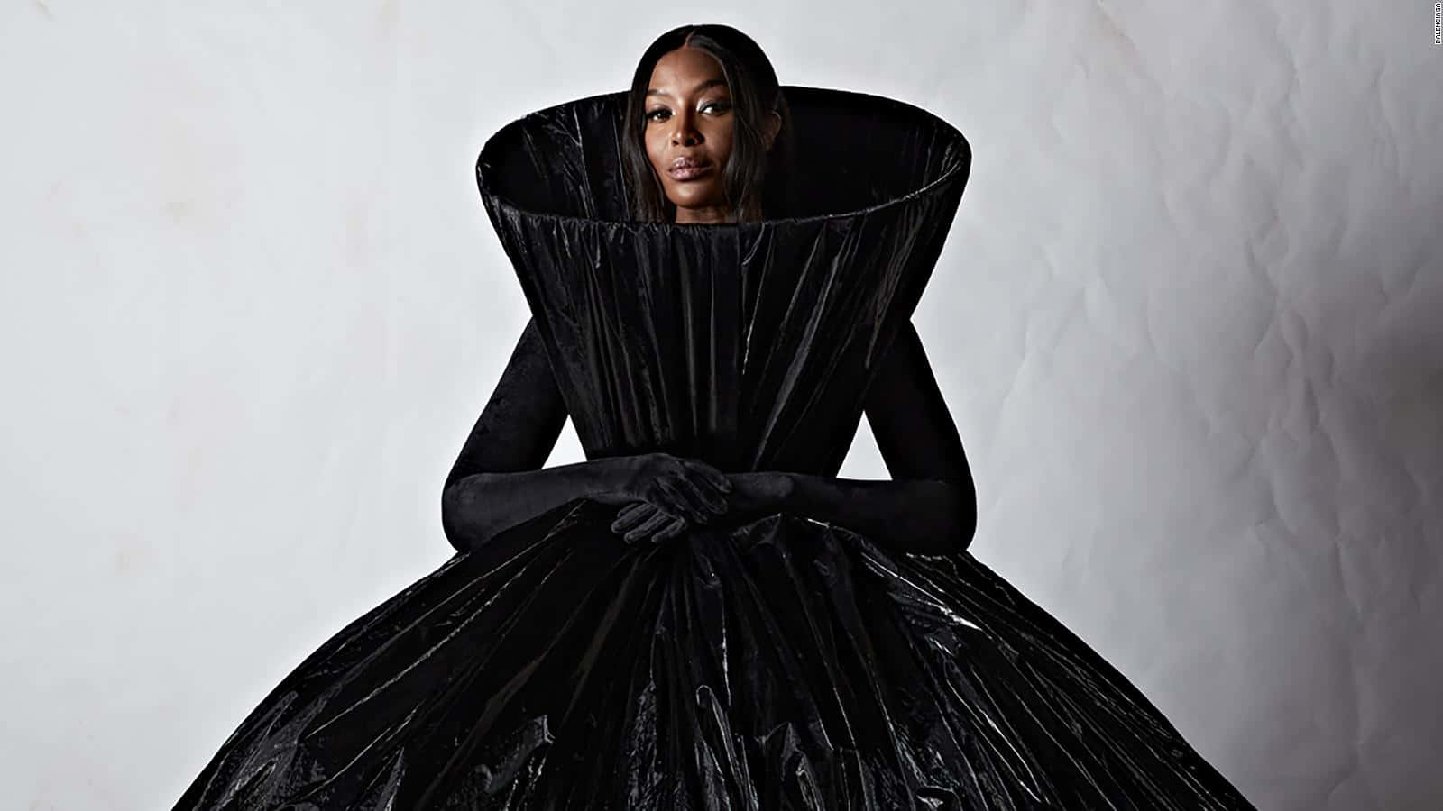 準備迎接新一季Balenciaga 壁紙 — 一位身穿戲劇性黑色塑膠長袍的女性，在帶有紋理的白色背景前擺姿勢，眼神強烈。