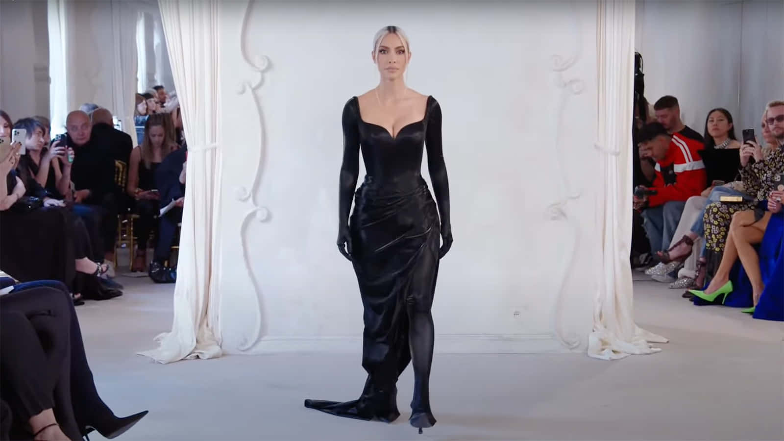 Balenciaga奢華高級時尚系列 壁紙 — 一位模特兒身穿時尚黑色緞面長裙，自信地走在簡約的白色伸展台上，兩旁坐著觀眾。