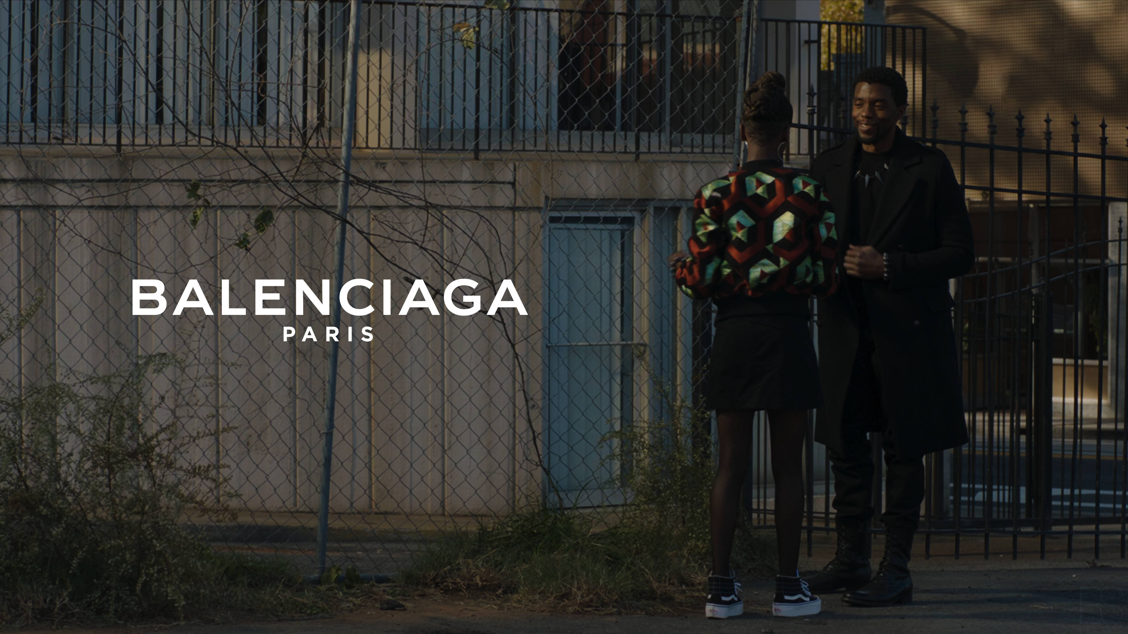 Balenciaga 巴黎封面照 壁紙 — 兩個穿著時尚的人站在鐵絲網圍欄附近，帶有「Balenciaga Paris」文字疊加在粗糙的城市環境中。