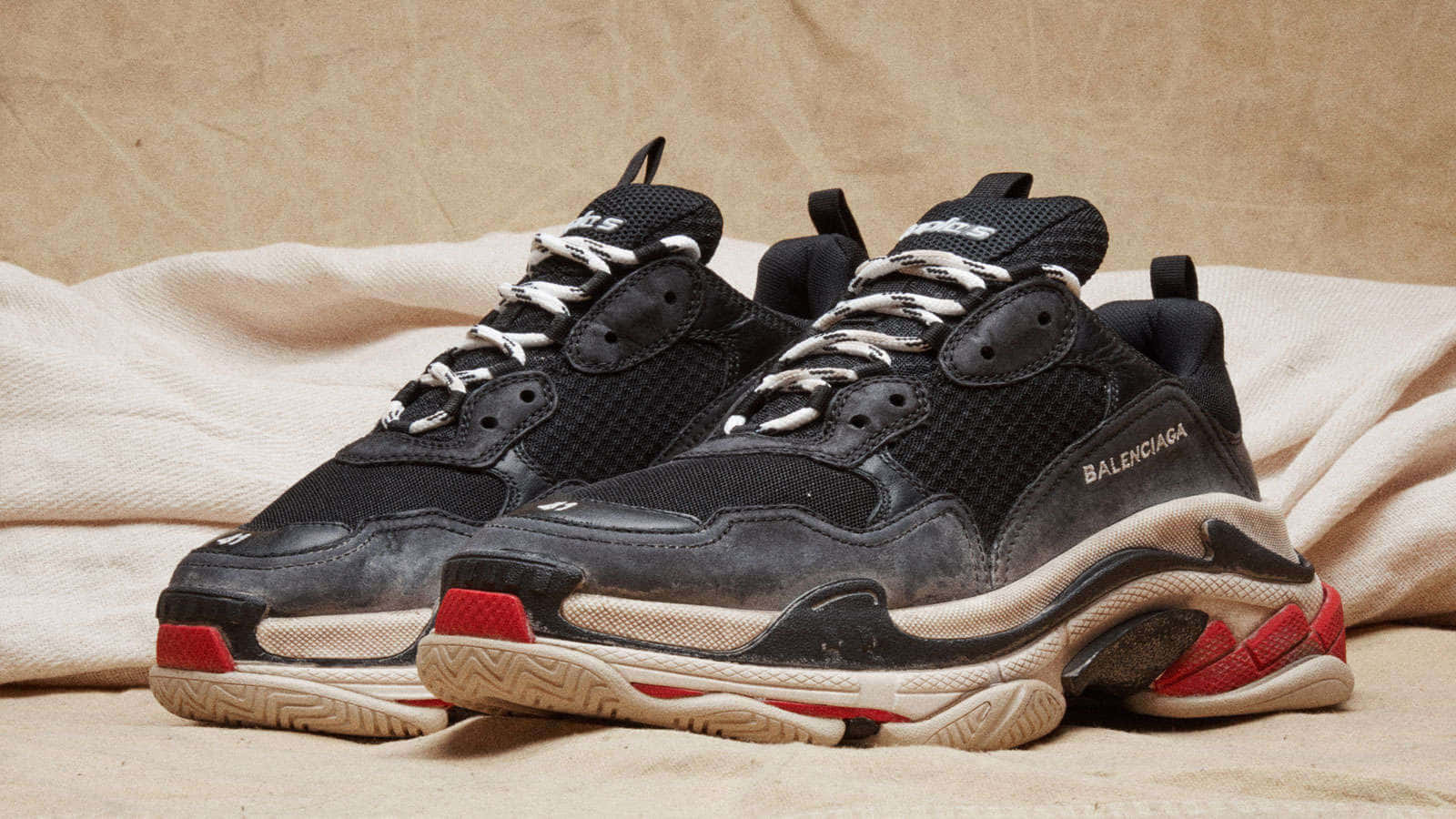 Balenciaga —— 時尚的重新定義 壁紙 — 一對帶有紅色點綴的黑色Balenciaga運動鞋，放在皺巴巴的米色布料上，背景有紋理。