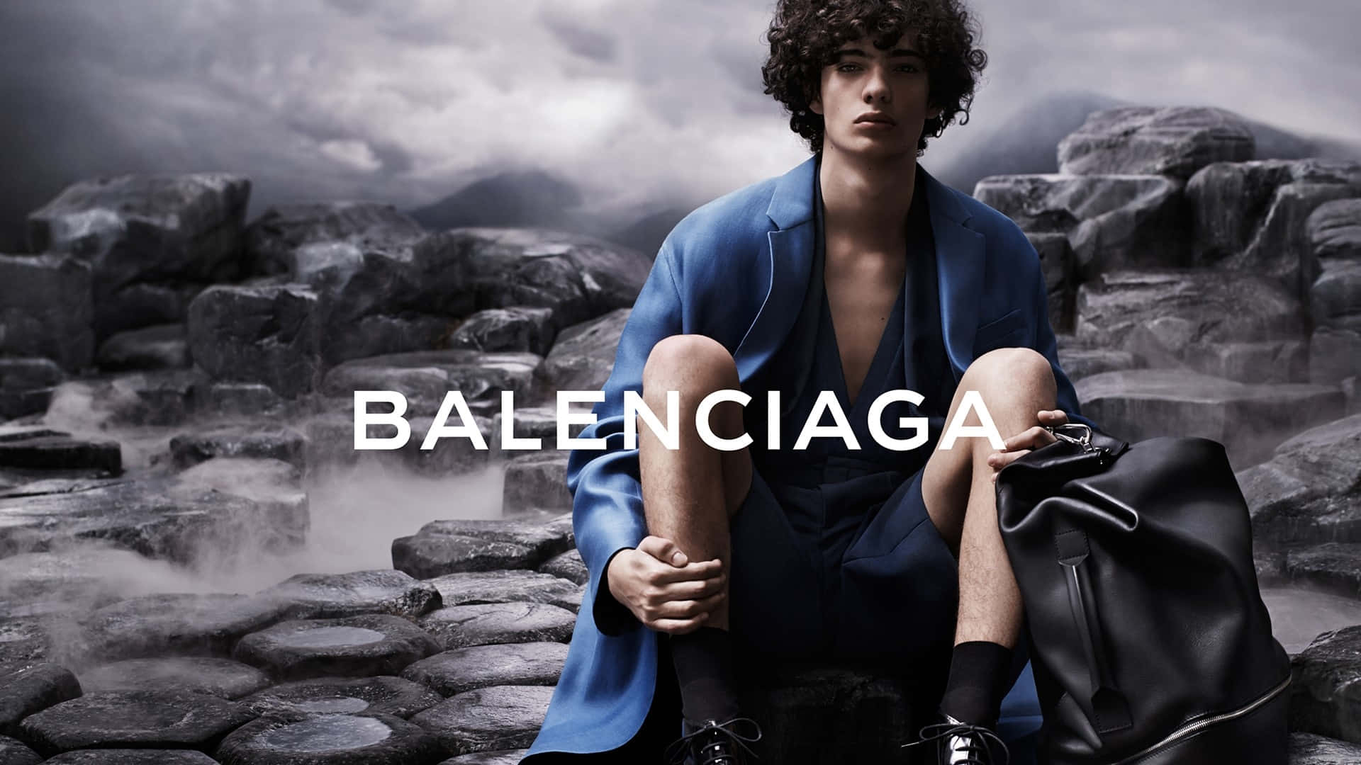 Balenciaga 標誌於優雅米色背景上 壁紙 — 一個捲髮男人坐在岩石地形上，穿著藍色西裝外套，拿著黑色袋子，在暴風雨的天空下。