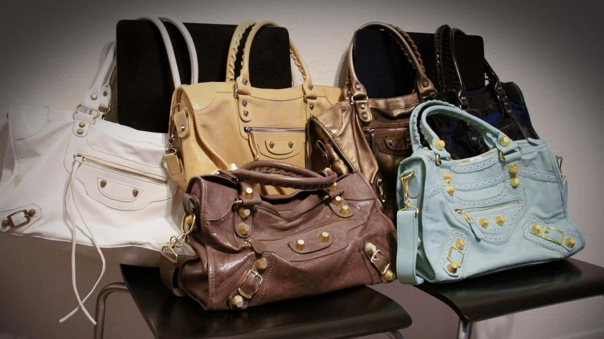 Balenciaga 的前衛時尚風采 壁紙 — 一系列奢華手袋，採用白色、米色、棕色和淺藍色皮革，配以金色五金件，排列在黑色凳子上，背景為中性牆壁。
