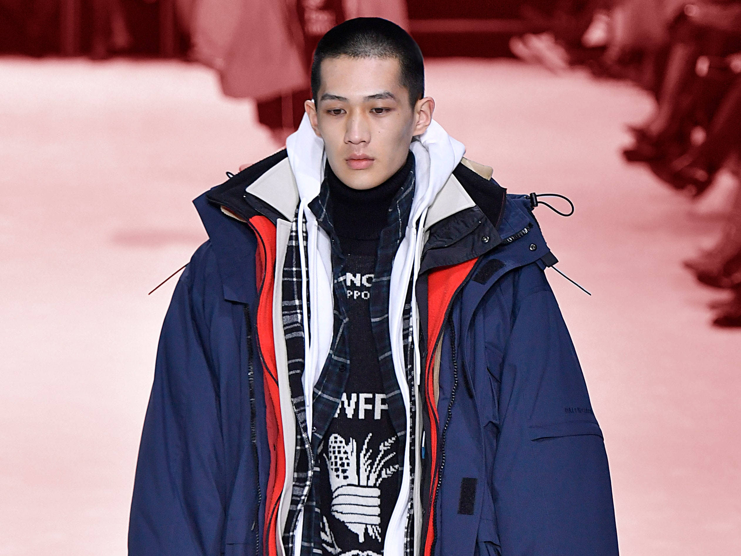 Balenciaga 藍色派克大衣 壁紙 — 一位模特兒在時裝天橋上走秀，穿著分層的冬季服裝，包括一件海軍藍色外套配紅色內裡和圖案高領毛衣。