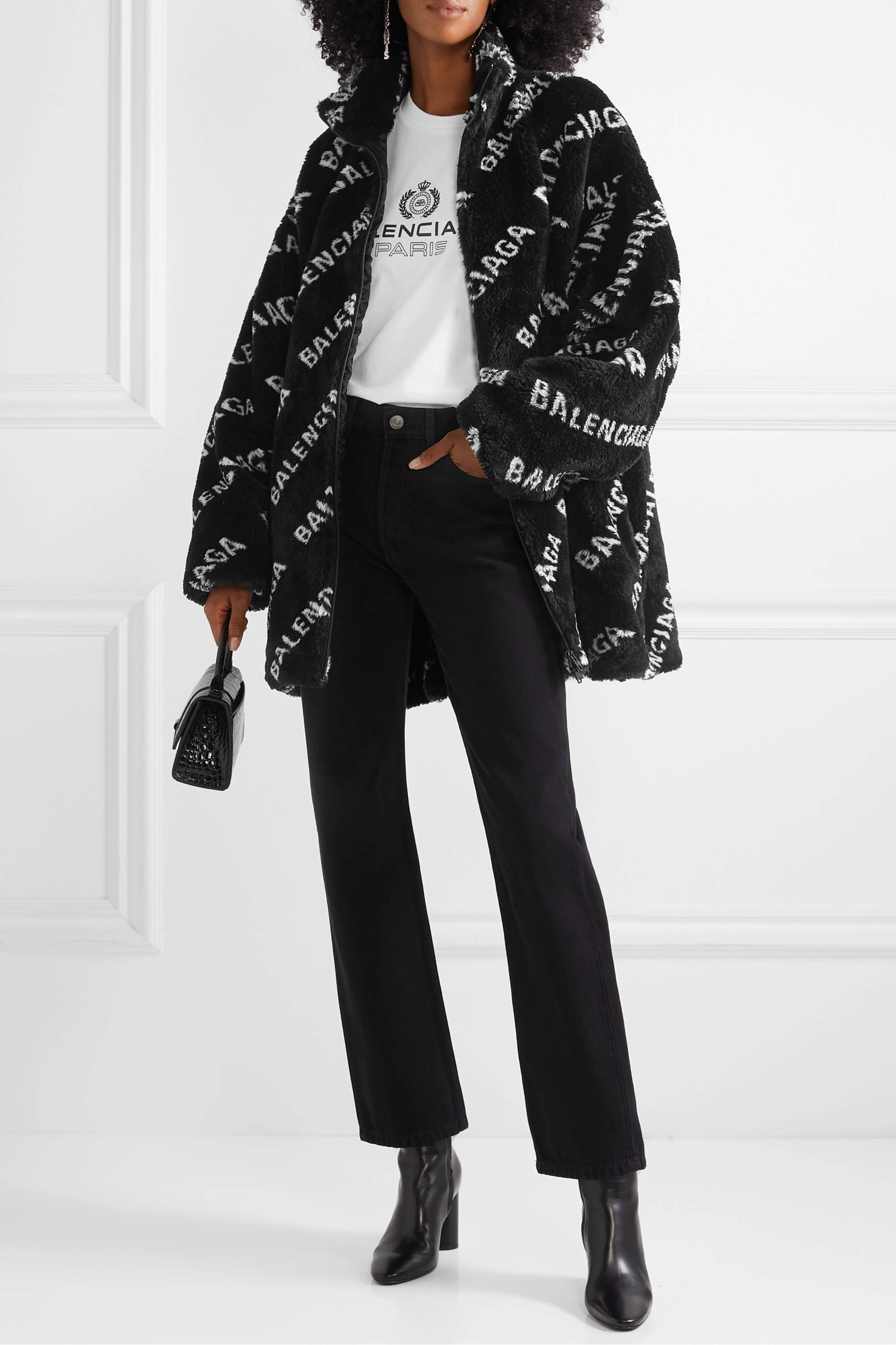 Balenciaga 黑色針織開衫 壁紙 — 一位時尚女性穿著黑色Balenciaga標誌印花泰迪外套，內搭白色T恤和黑色褲子，背景是簡約的白色牆壁。