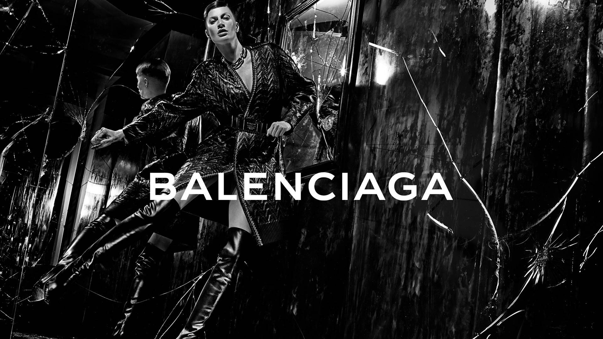 Balenciaga單色黑 壁紙 — 一位模特兒穿著質感外套和過膝靴，在破碎玻璃背景前，於高對比黑白Balenciaga廣告中戲劇性地擺姿勢。