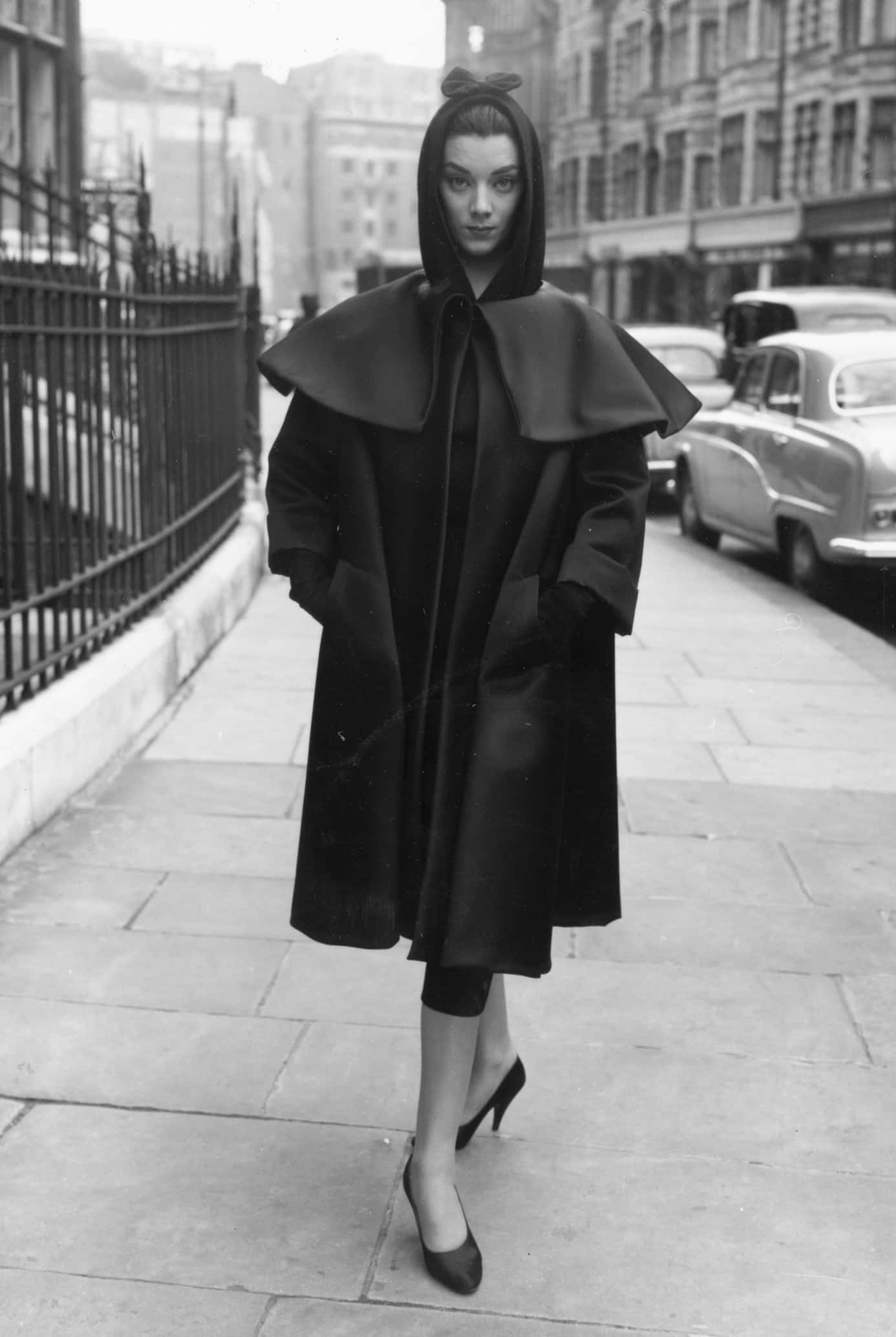 Balenciaga服裝的休閒優雅 壁紙 — 一位身穿戲劇性黑色連帽外套的女性自信地走在城市人行道上，經過復古汽車和鐵柵欄。
