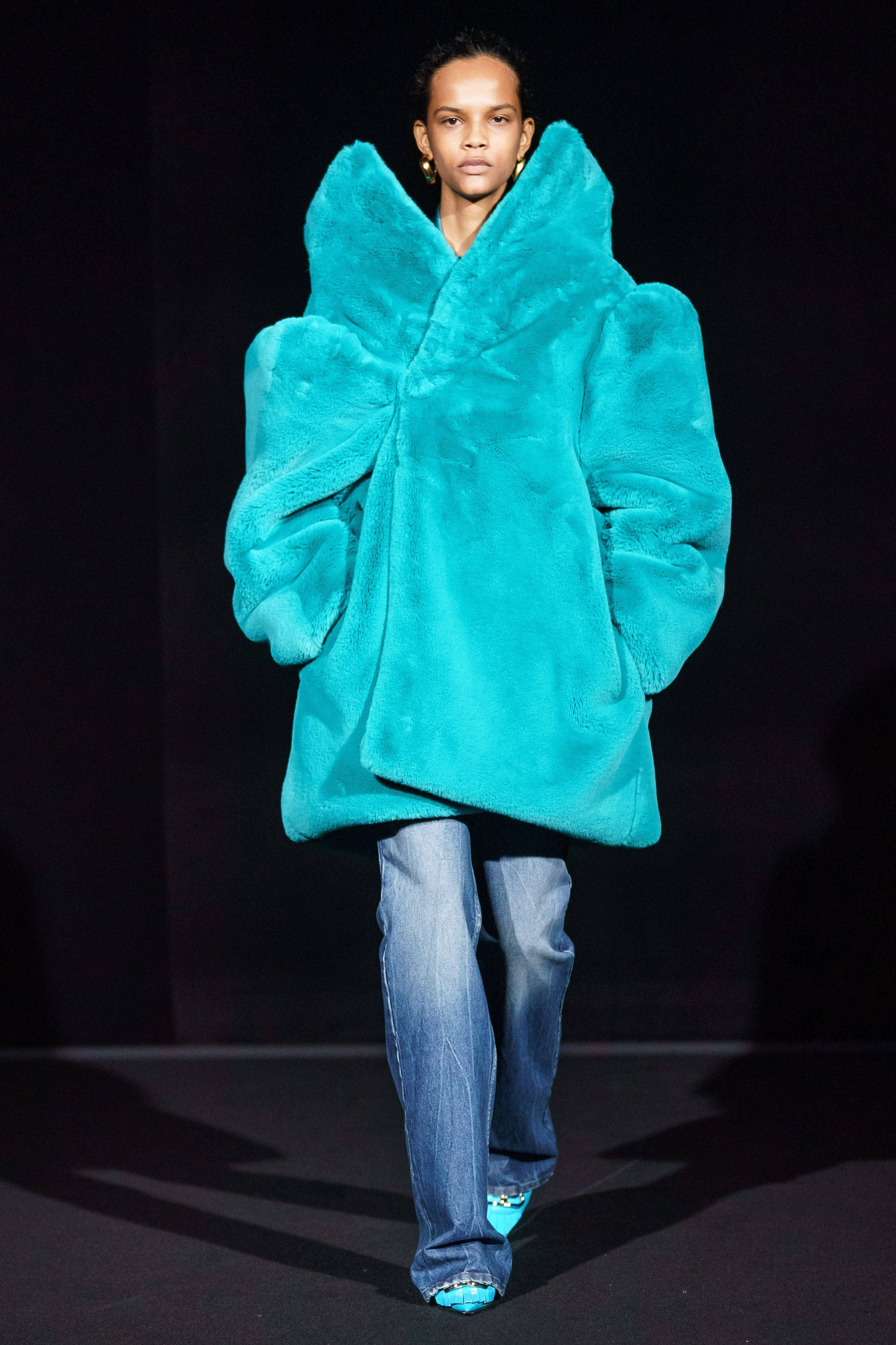Balenciaga藍色絨毛大衣 壁紙 — 模特兒在舞台燈光下自信地穿著一件寬大的青綠色人造毛皮大衣和藍色牛仔褲。