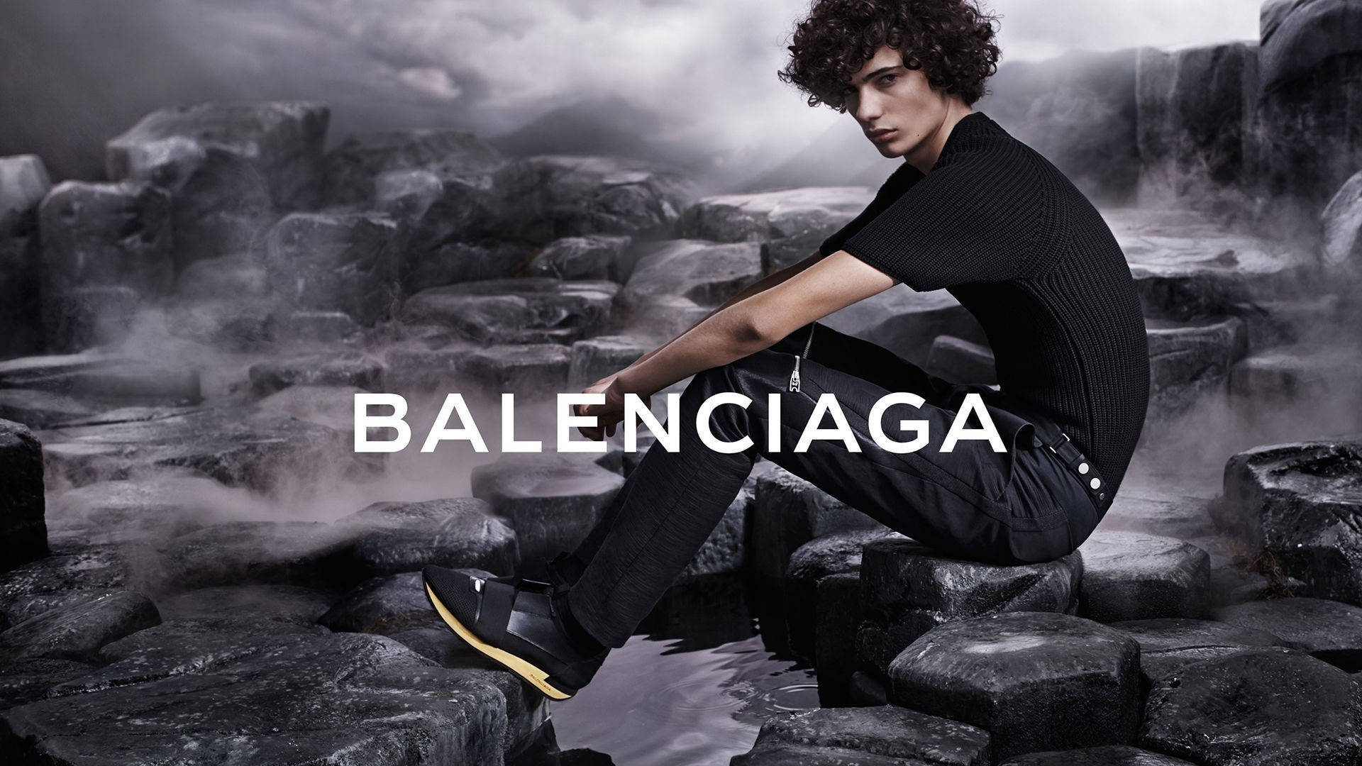 Balenciaga黑色襯衫 壁紙 — 一位捲髮年輕男子坐在霧氣瀰漫的火山岩上，身穿黑色 Balenciaga 服裝和黃色鞋底的鞋子。