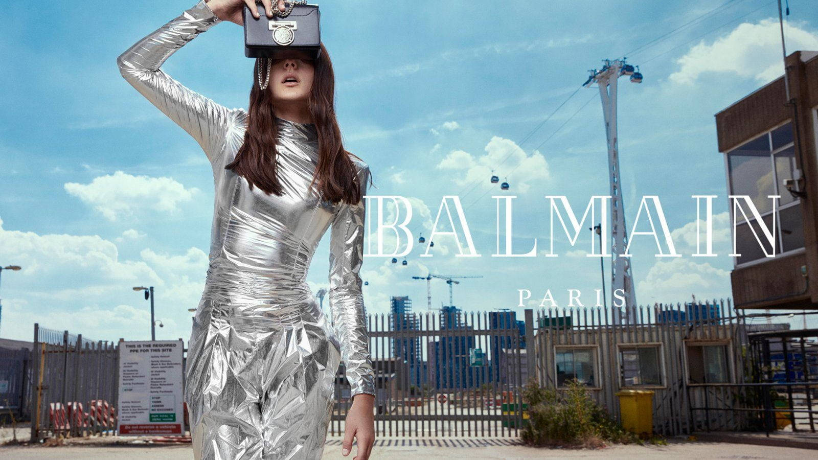 銀色 Balmain 套裝 壁紙 — 一位身穿閃亮銀色連衣裙的女性，在藍天白雲和建築工地背景下，將黑色手提包舉過眼睛。