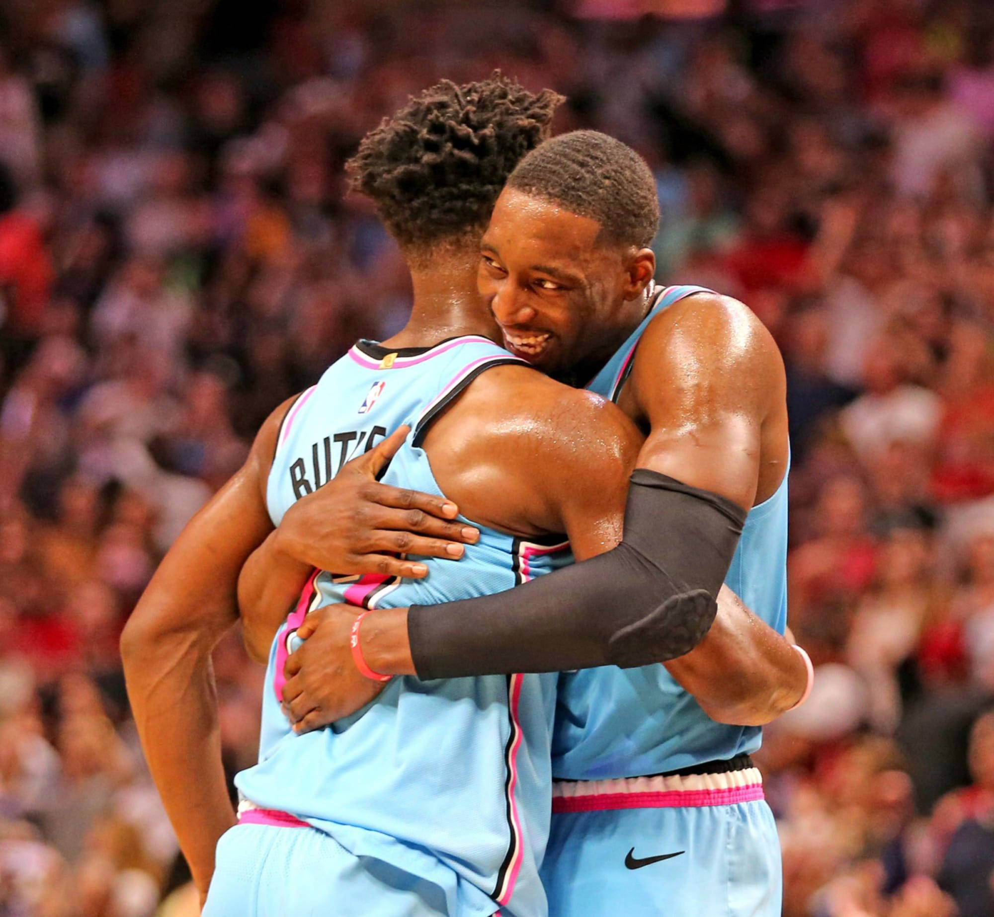 Bam Adebayo 與吉米·巴特勒 壁紙 — 兩名身穿淺藍色球衣的籃球運動員在球場上歡樂地擁抱，身後是模糊的人群。