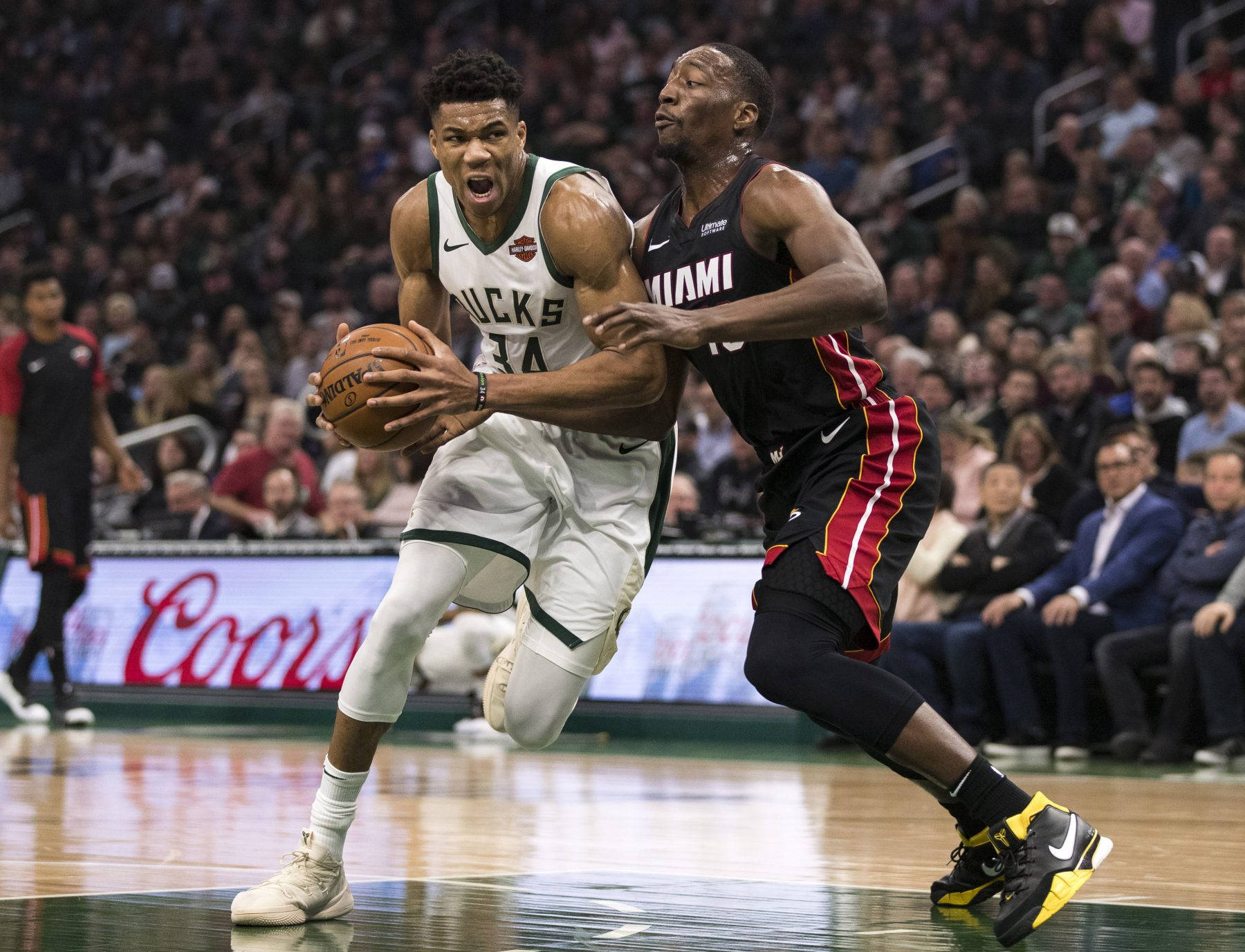 Bam Adebayo 防守 Giannis Antetokounmpo 壁紙 — 一名身穿白色公鹿球衣的籃球運動員在激烈的比賽中，在座無虛席的球場背景下，帶球突破邁阿密熱火隊的防守球員。