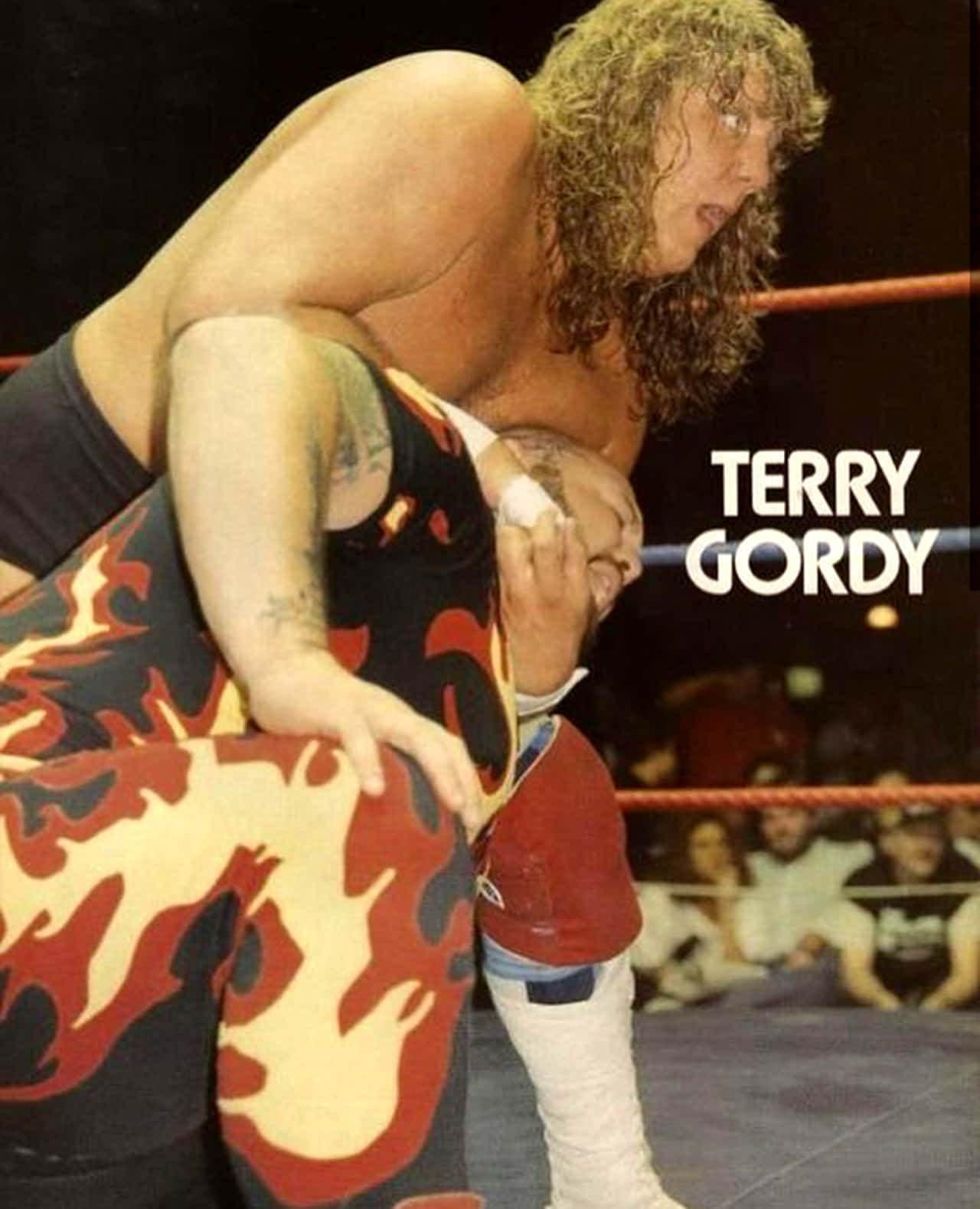 Bam Bam Bigelow VS. Terry Gordy 80年代經典WWE遊戲場景 壁紙 — Terry Gordy 在明亮的場館燈光下，穿著火焰圖案緊身衣，在激烈的摔角比賽中舉起對手。