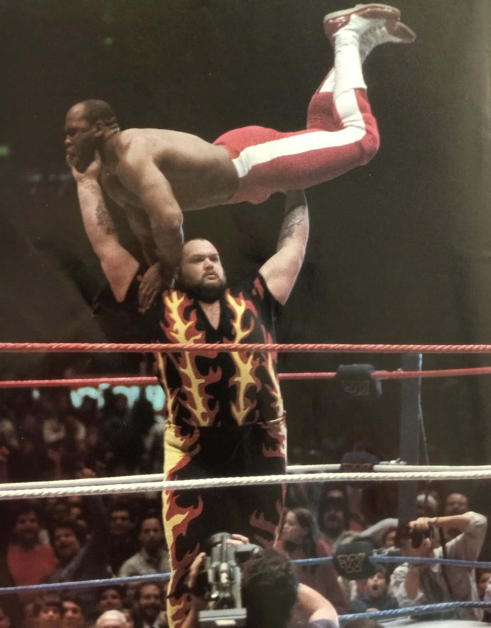 Bam Bam Bigelow 對決 Virgil 1988 壁紙 — 一名穿著火焰圖案短褲的摔角手在明亮的場館燈光下，在摔角台上將另一名摔角手高舉過頭。
